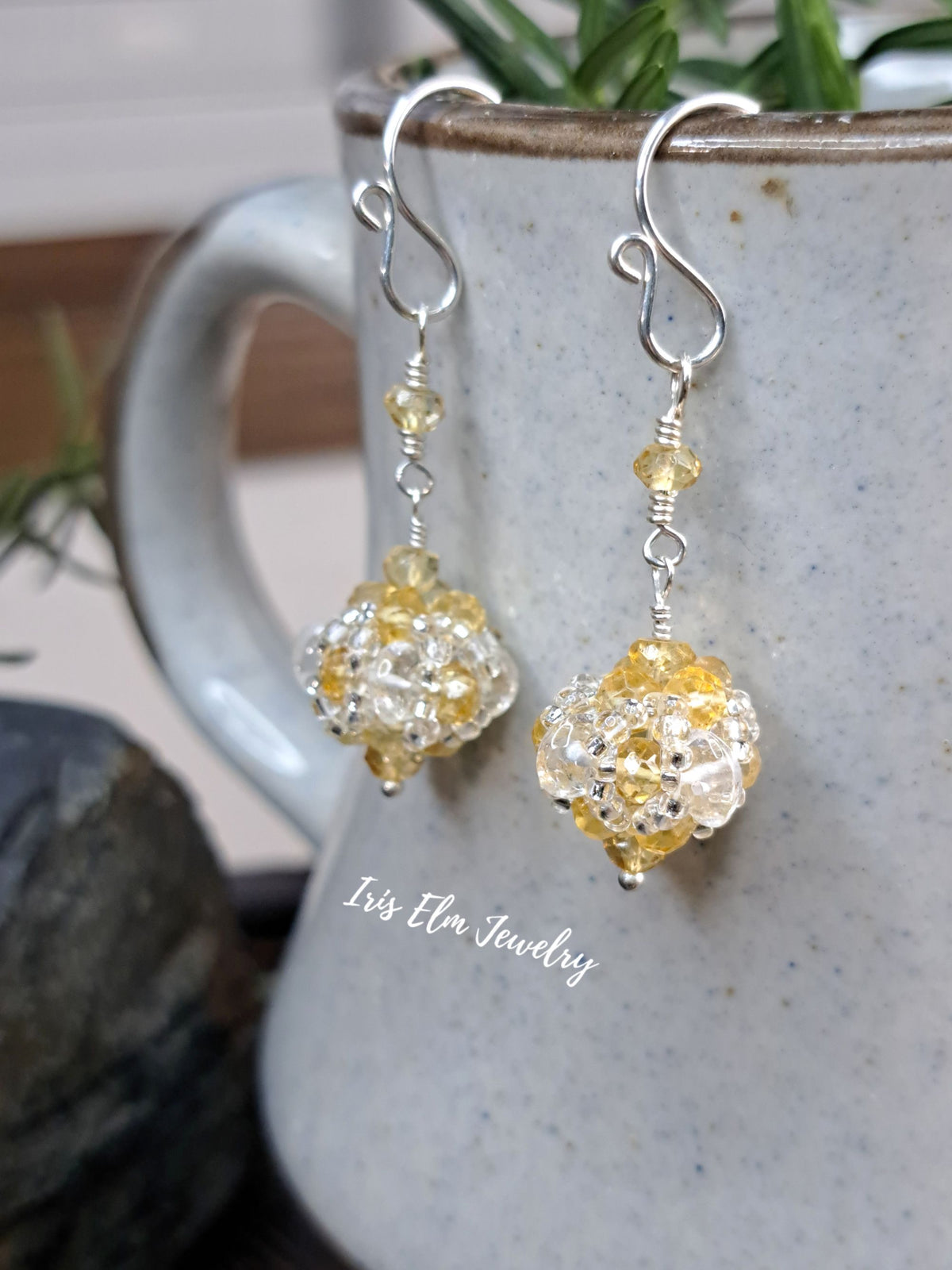 A pair of handmade dangle earrings with clear quartz and yellow citrine faceted beads and silver hooks, hanging in front of a speckled gray ceramic mug. The brand name 'Iris Elm Jewelry' is watermarked on the mug.
