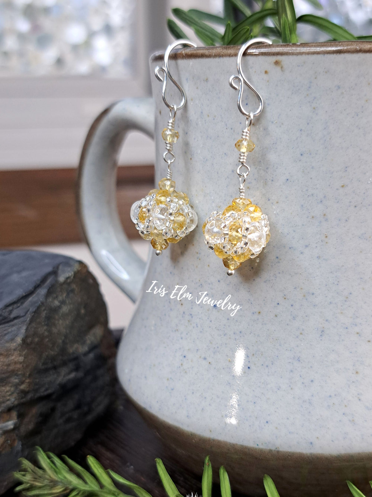 A pair of handmade dangle earrings with clear and yellow beads and silver hooks, hanging from the rim of a speckled ceramic mug. The mug is styled with pine branches and a dark stone on a wooden surface.