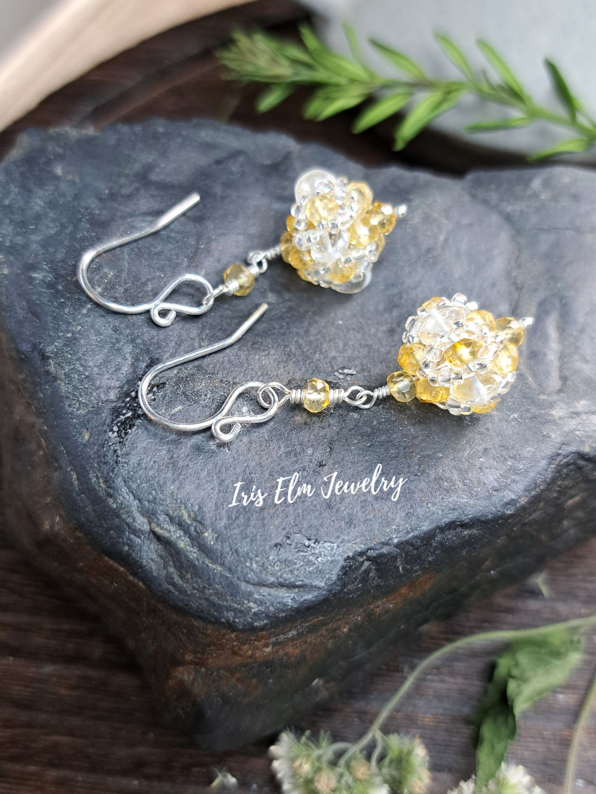 A pair of handmade dangle earrings with silver fishhook wires and textured, beaded floral clusters in white and translucent yellow, displayed on a dark slate stone with the brand name 'Iris Elm Jewelry' printed on it.