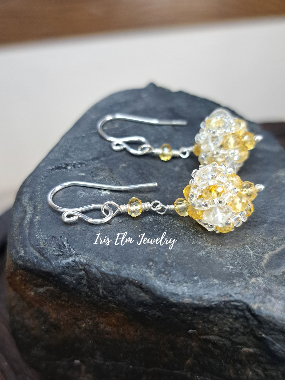A pair of handmade dangle earrings with silver fishhook wires and clear beads wrapped in yellow glitter, displayed on a dark textured rock. The brand name 'Iris Elm Jewelry' is printed on the rock.