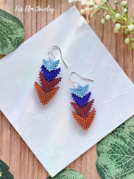 Colorful beaded blue and burnt orange tiered triangle seed bead earrings on a white napkin with green leaves and flowers in the background.