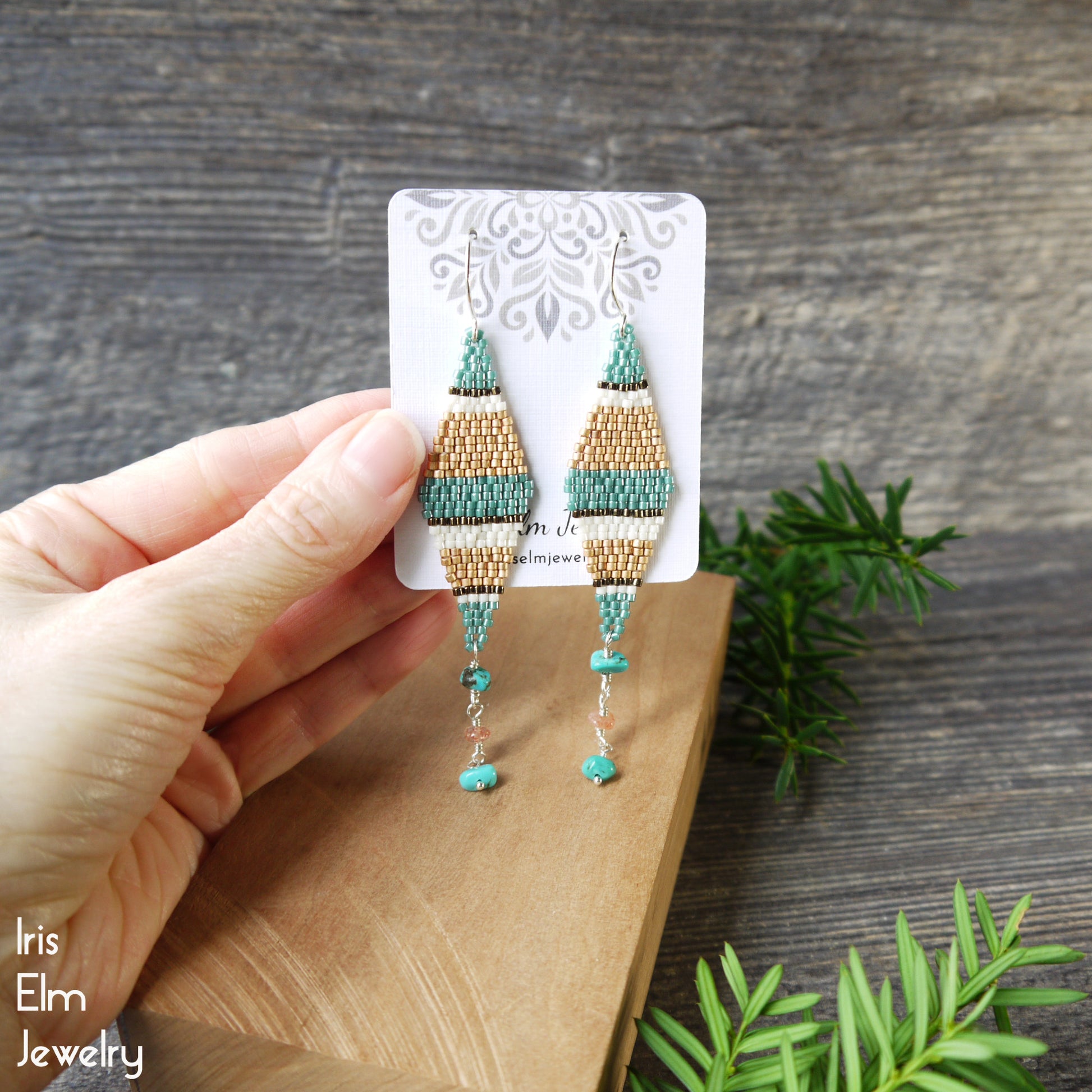 Hand holding a pair of beaded seed bead kite and turquoise and sunstone earrings on a wooden surface with greenery