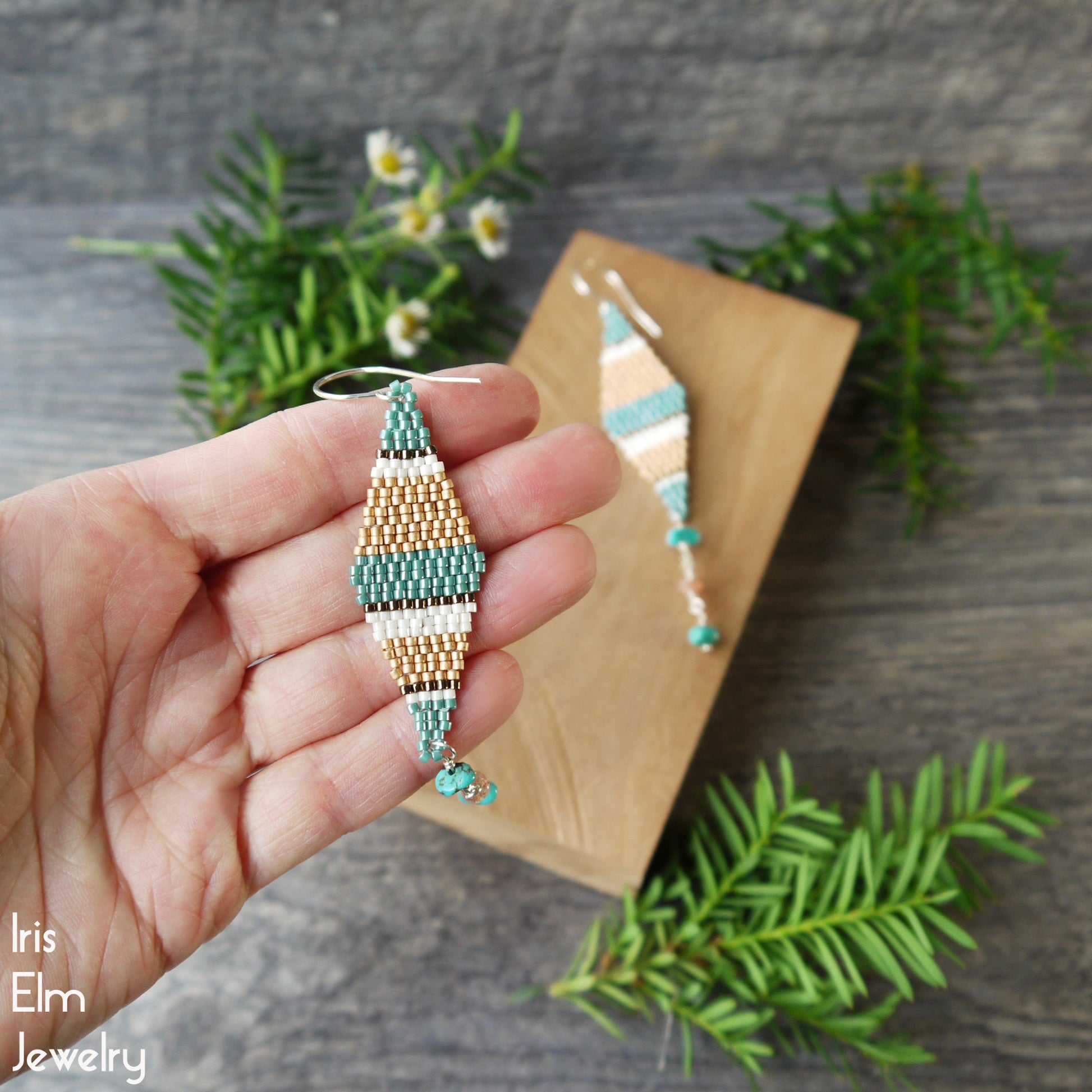Hand holding a beaded seed bead kite earring with blue and copper colors, placed on a wooden block with plants in the background.