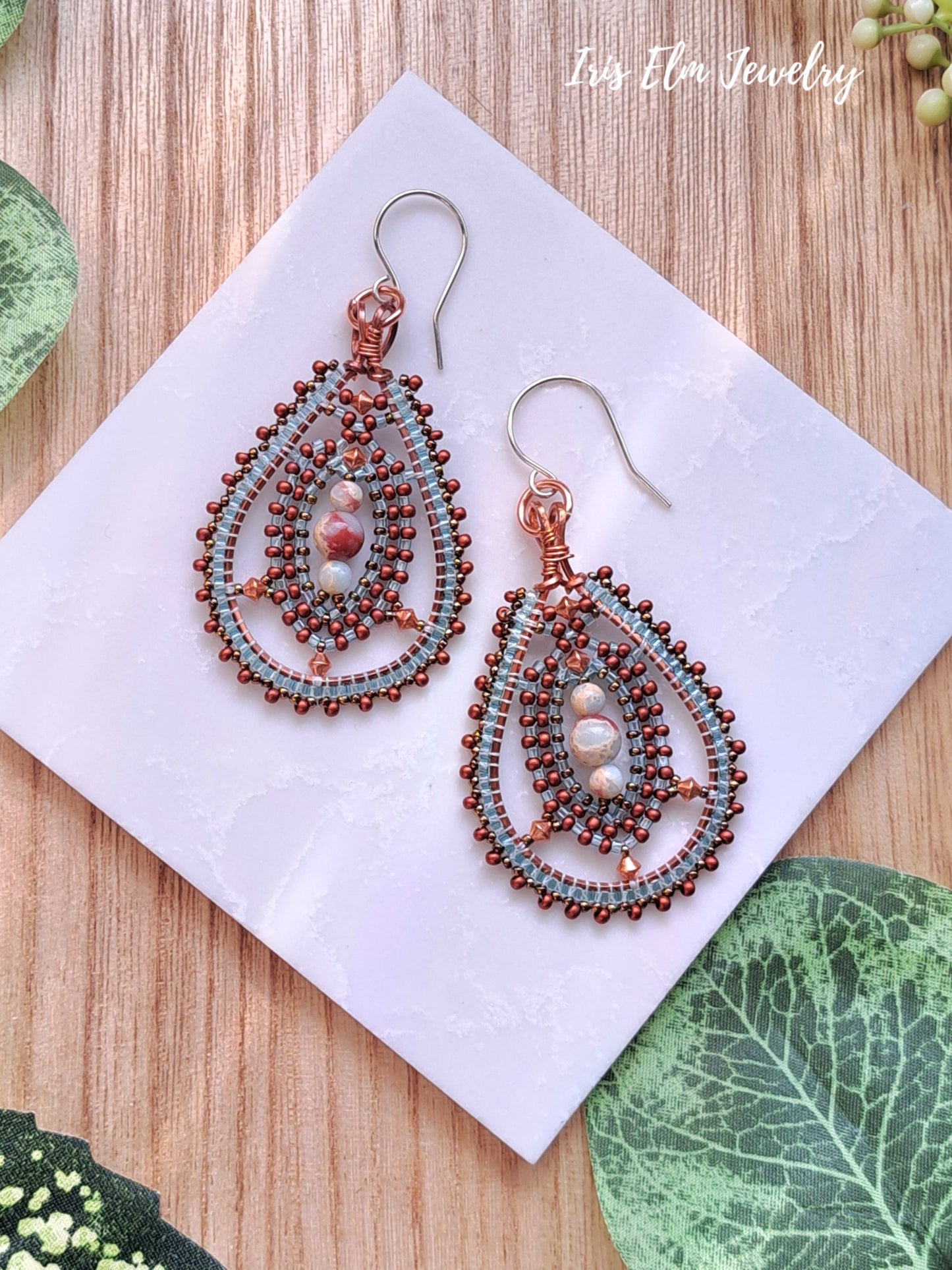 Pair of beadwork impression jasper and seed bead teardrop copper earrings on a white card with green leaves in the background