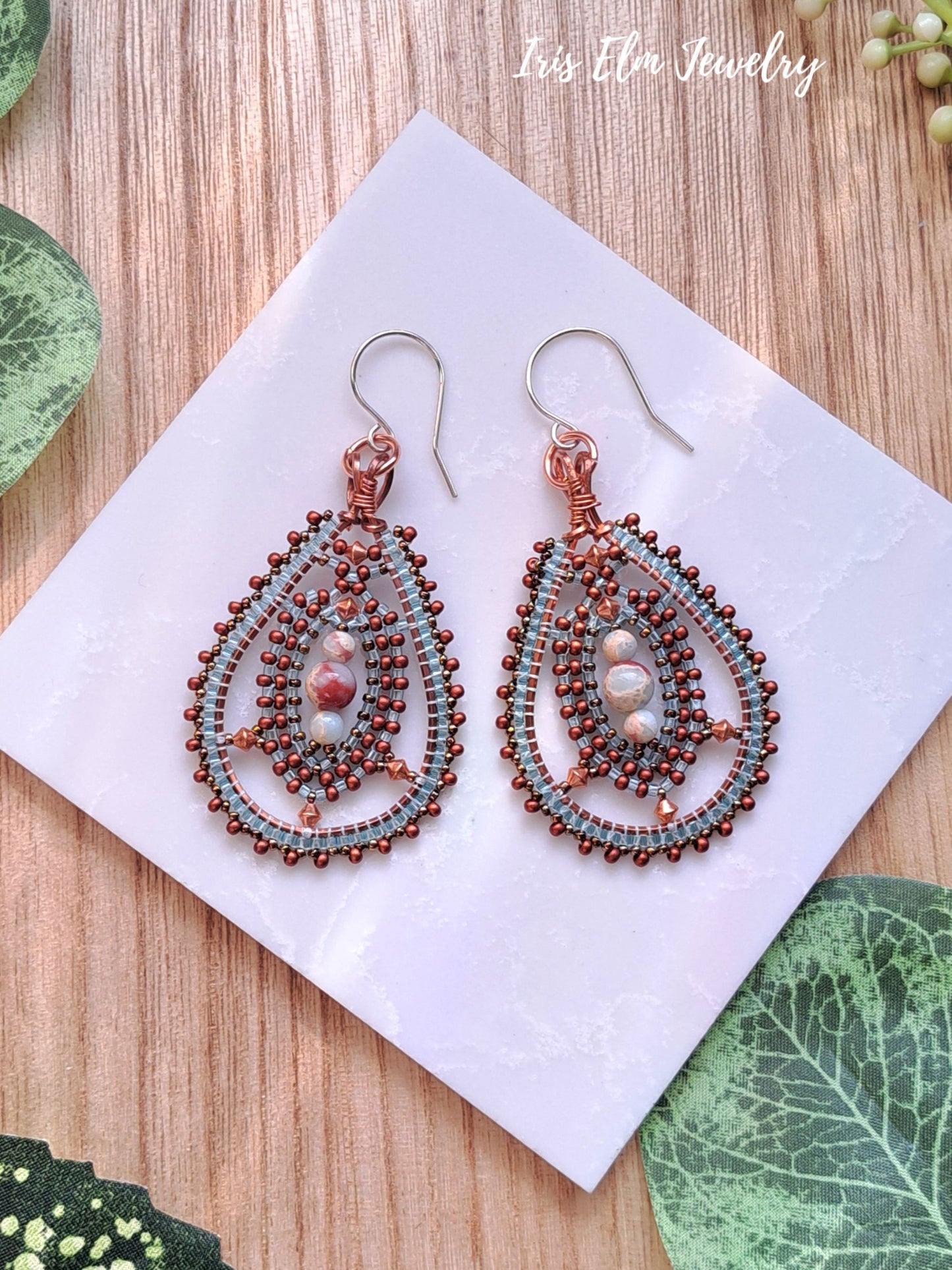 Pair of beaded jasper copper seed bead stone teardrop statement earrings on a white card with green leaves around, branded 'Iris Elm Jewelry'.