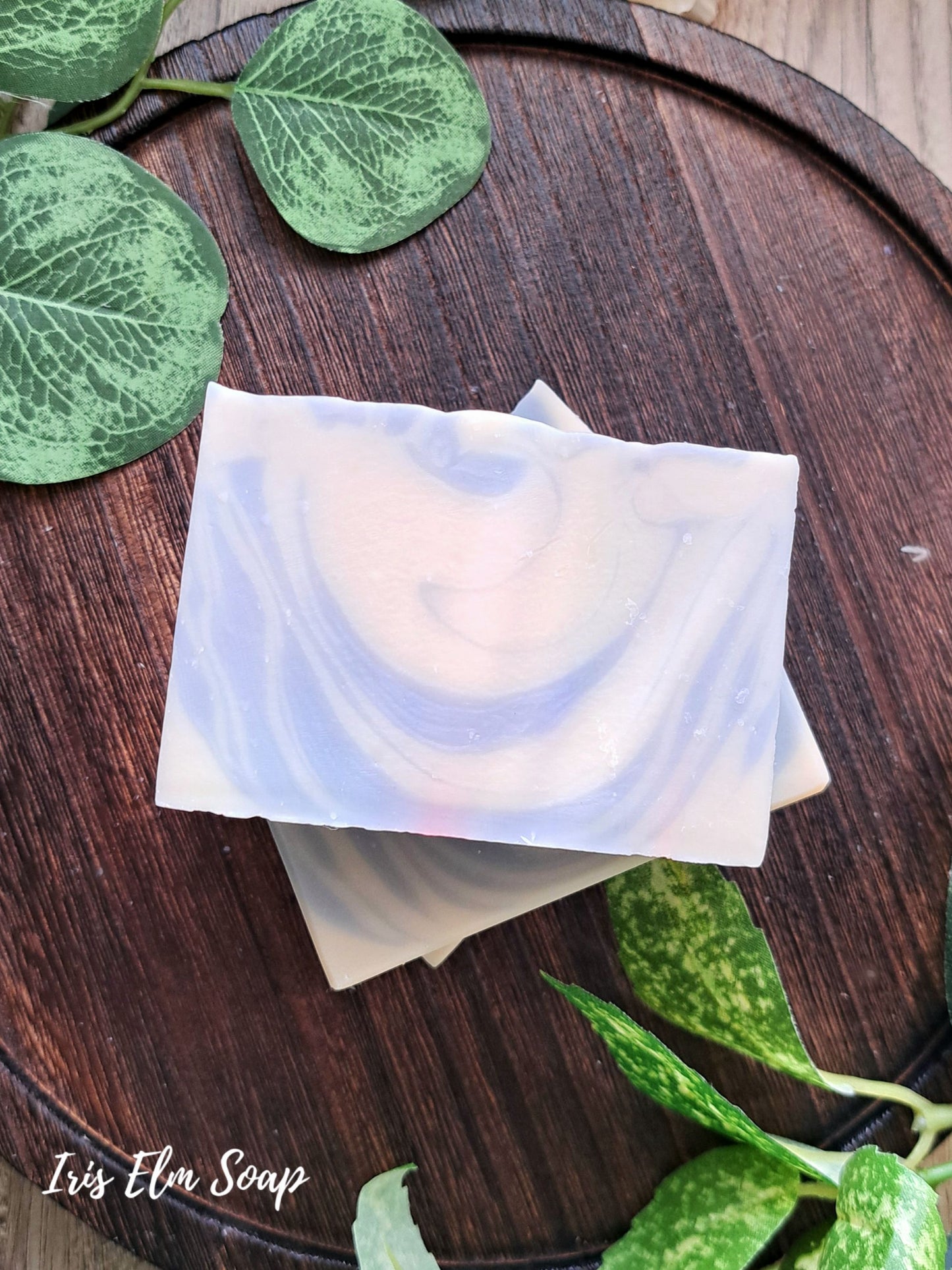 Two pieces of clean cotton handmade soap with a swirling pattern on a wooden surface with green leaves.