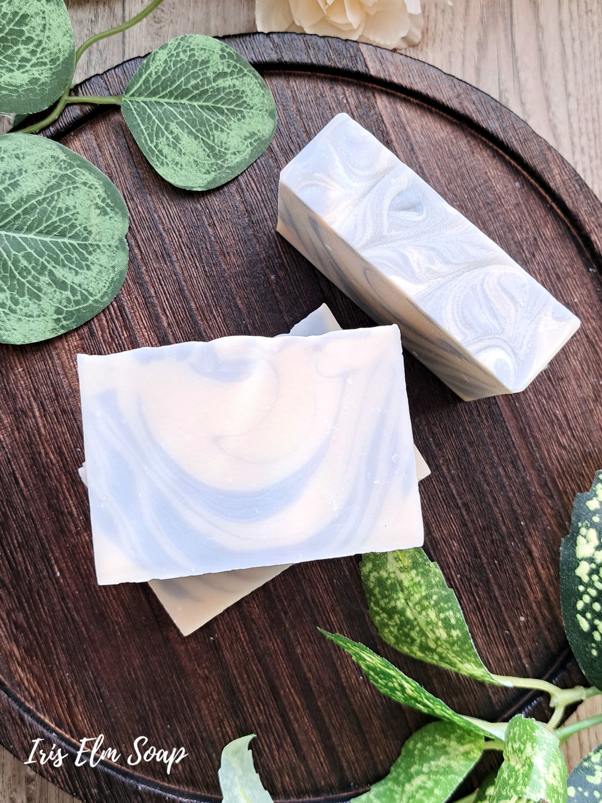 Two bars of clean cotton handmade soap on a wooden surface with green leaves around