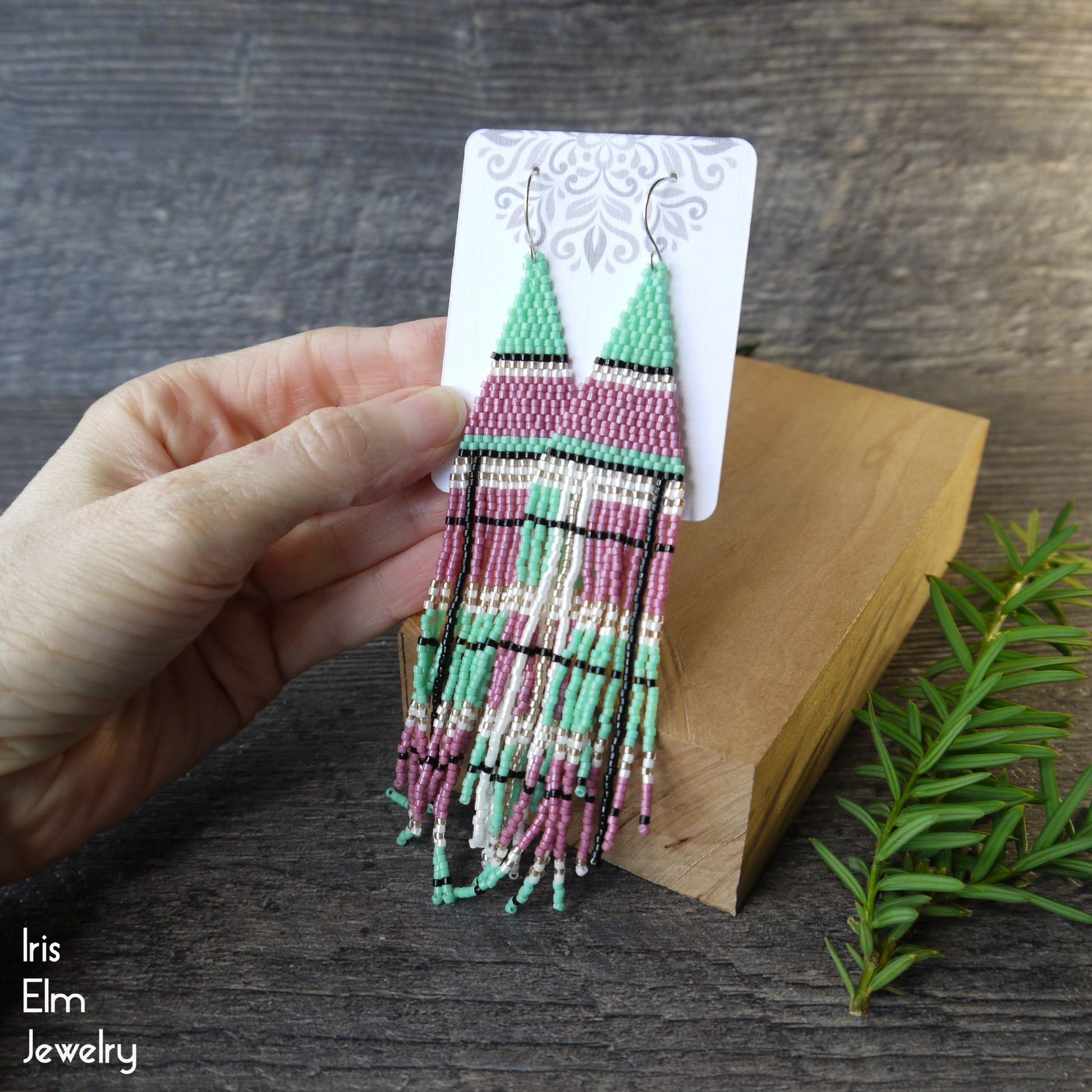 Hand holding a pair of beaded seed bead plaid pink and green fringe earrings on a wooden block with a natural background