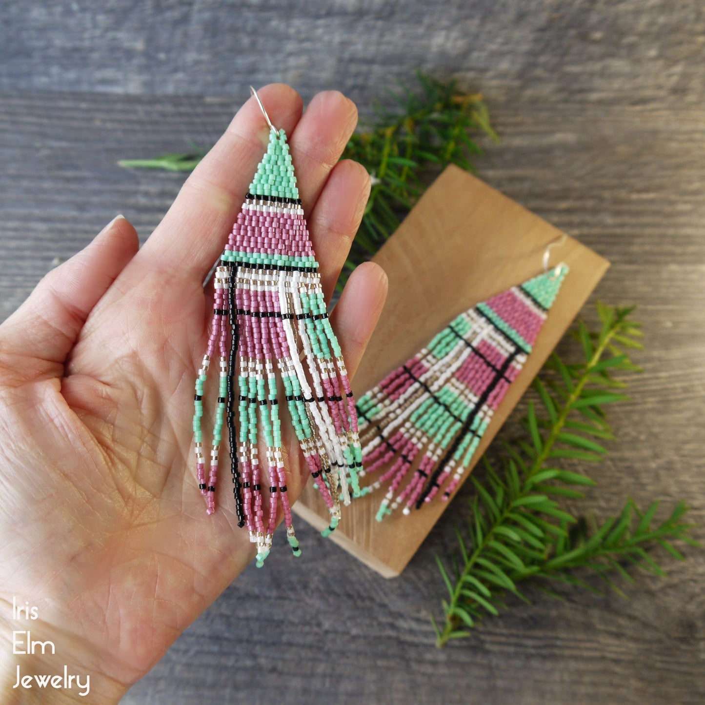 Beaded pink and green plaid fringe earrings held in a hand with a wooden surface and greenery in the background