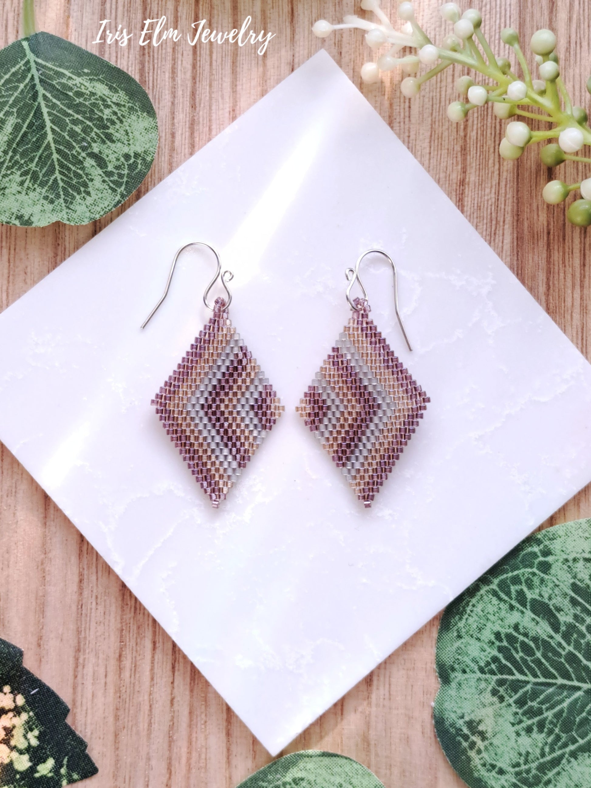 Pair of diamond-shaped chevron pink golden and grey seed bead earrings on a white card with green leaves and flowers in the background.