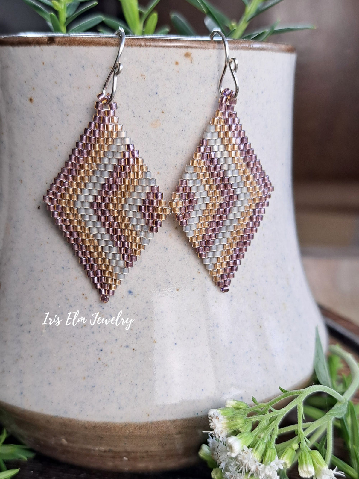 A pair of handmade beaded dangle earrings with a geometric diamond pattern in silver, gold, and dusty rose, hanging from silver fishhook wires. They are displayed on a rustic beige ceramic pot with green sprigs.