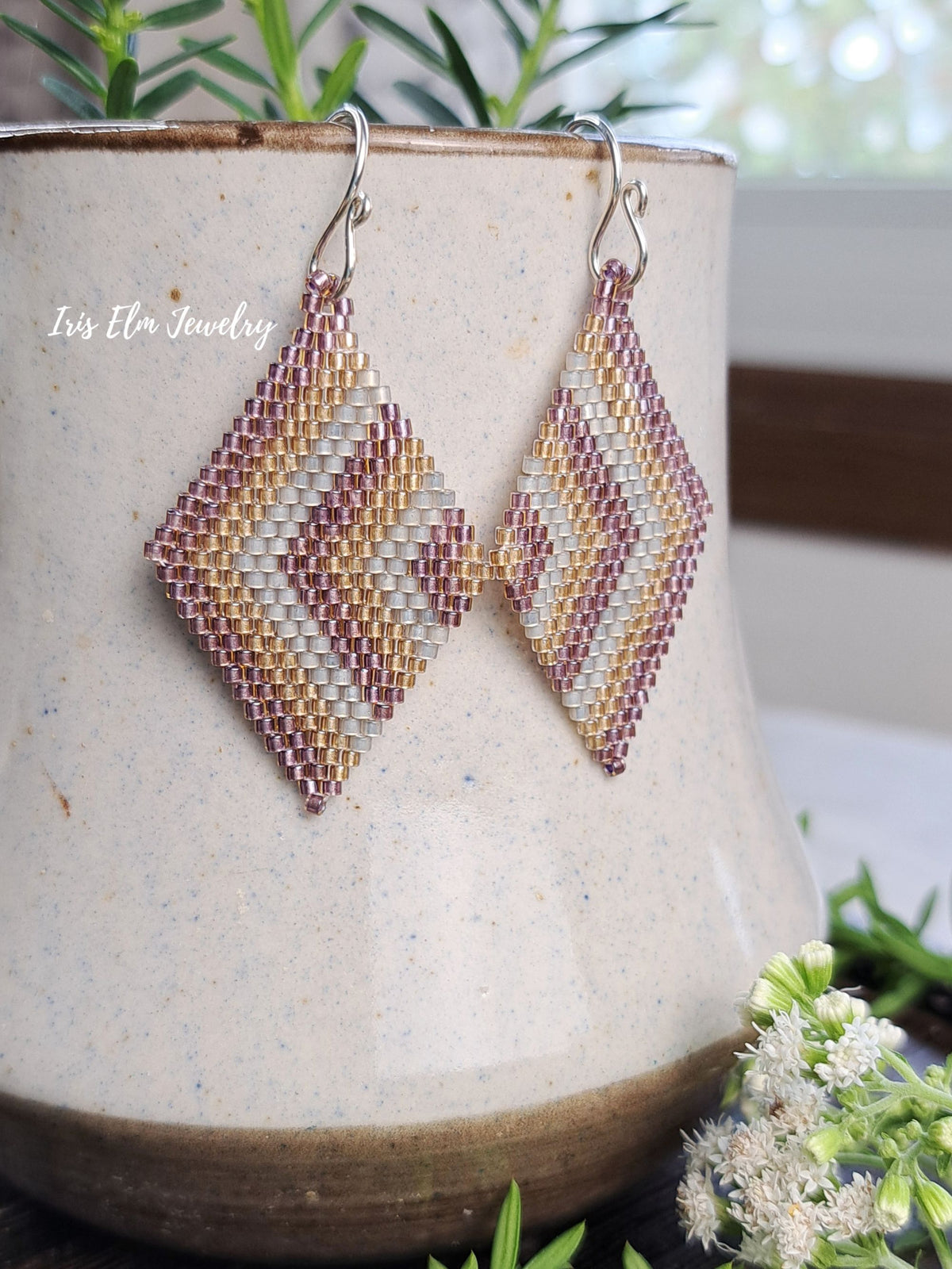 A pair of handmade beaded dangle earrings with a geometric diamond pattern in cream, gold, and dusty rose, hanging from silver fishhook wires. They are displayed on a speckled beige ceramic pot with green foliage and small white flowers in the background.