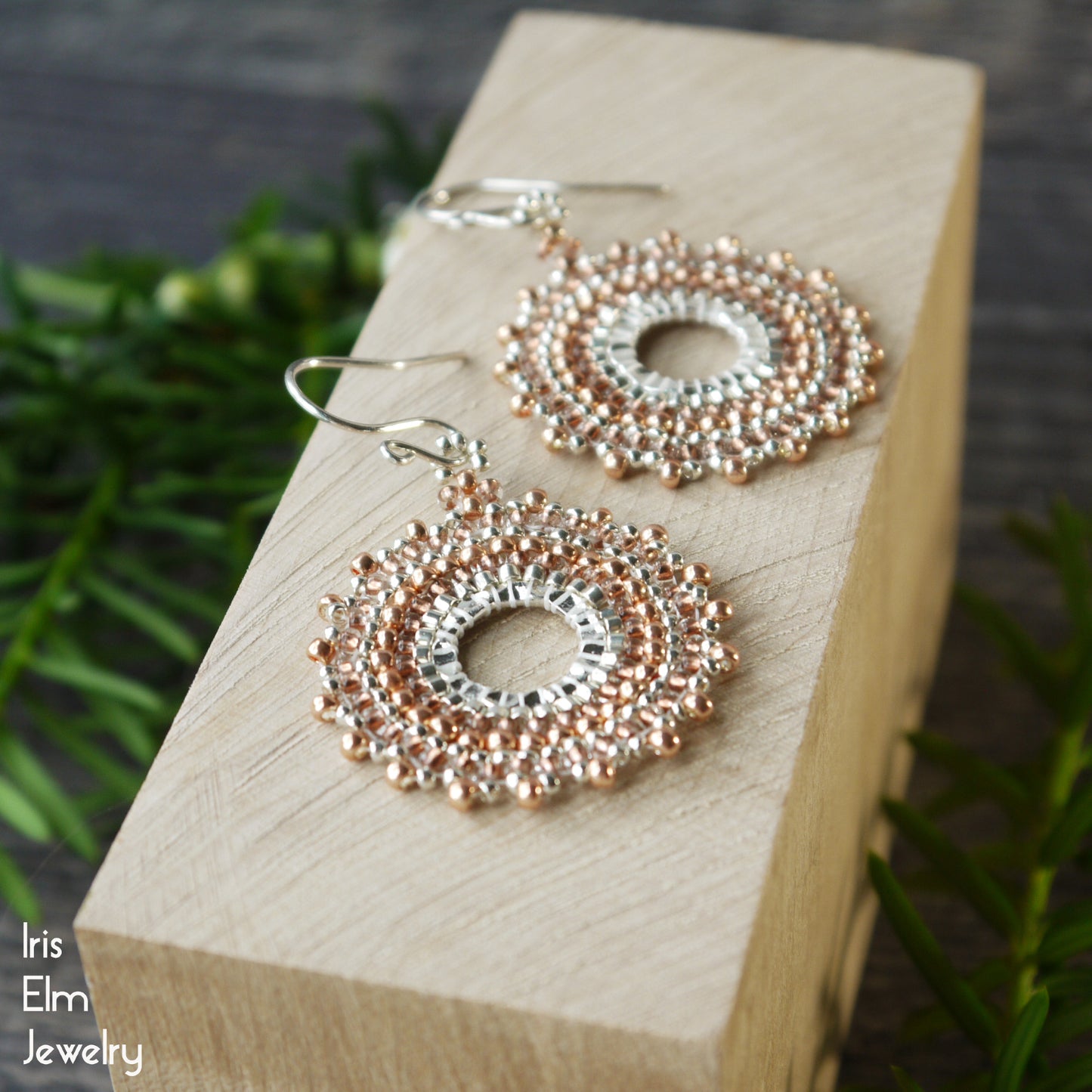 Pair of handmade beaded copper and silver woven seed bead earrings on a wooden block with a blurred natural background