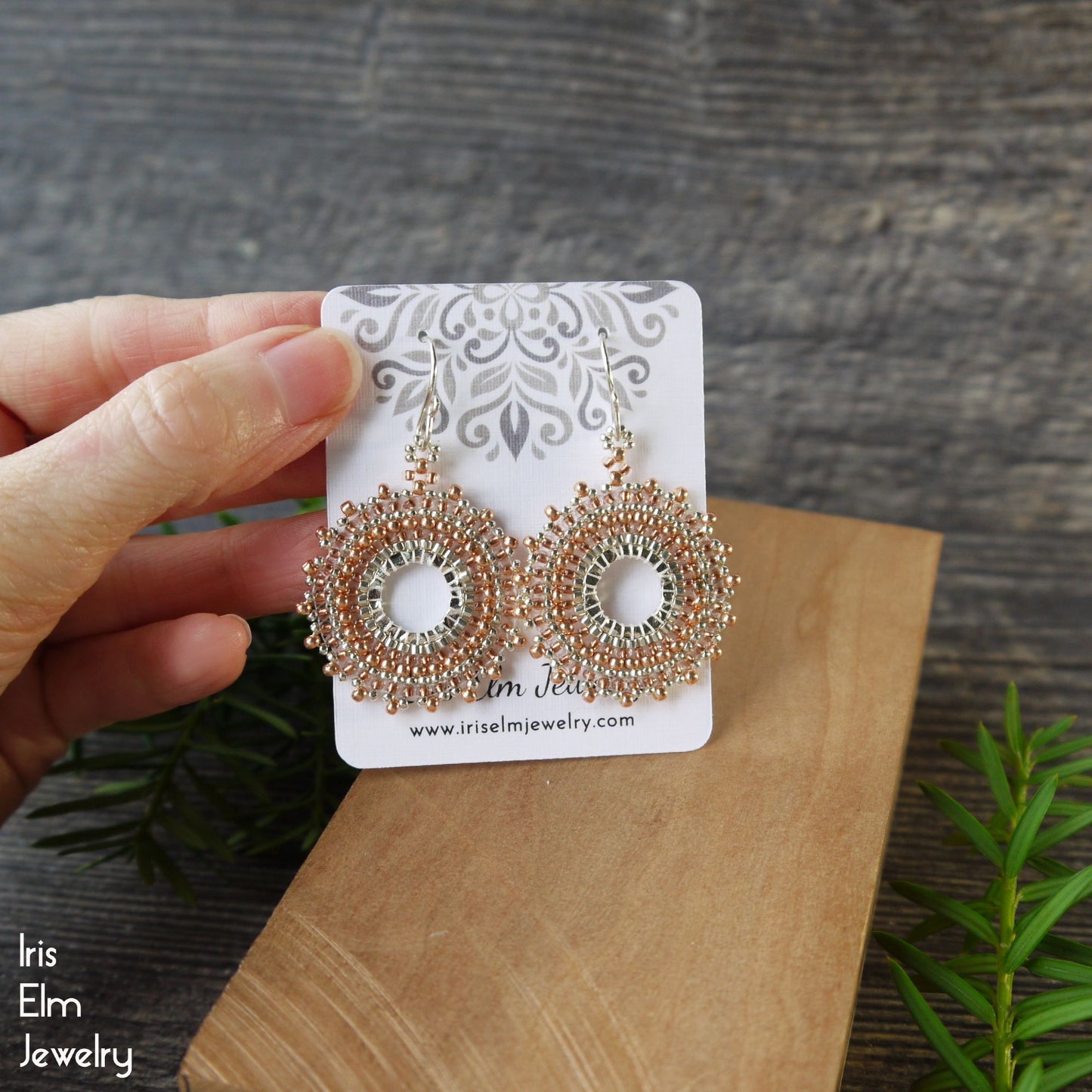 Copper hoop earrings on a 'Iris Elm Jewelry' branded card held by a hand with a wooden surface and plant in the background.