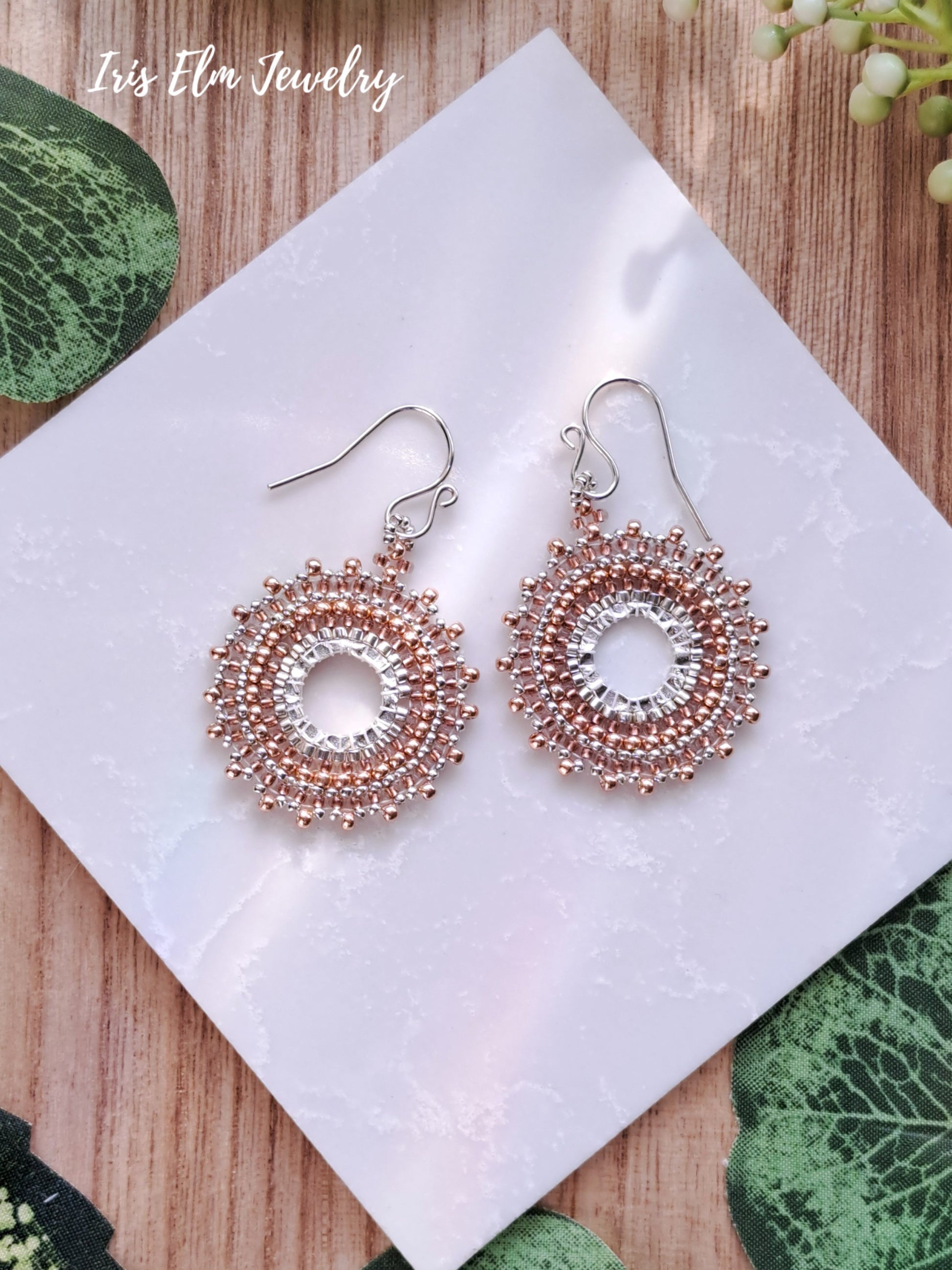 Pair of beaded copper seed bead woven hoop earrings on a white tile with 'Iris Elm Jewelry' branding.