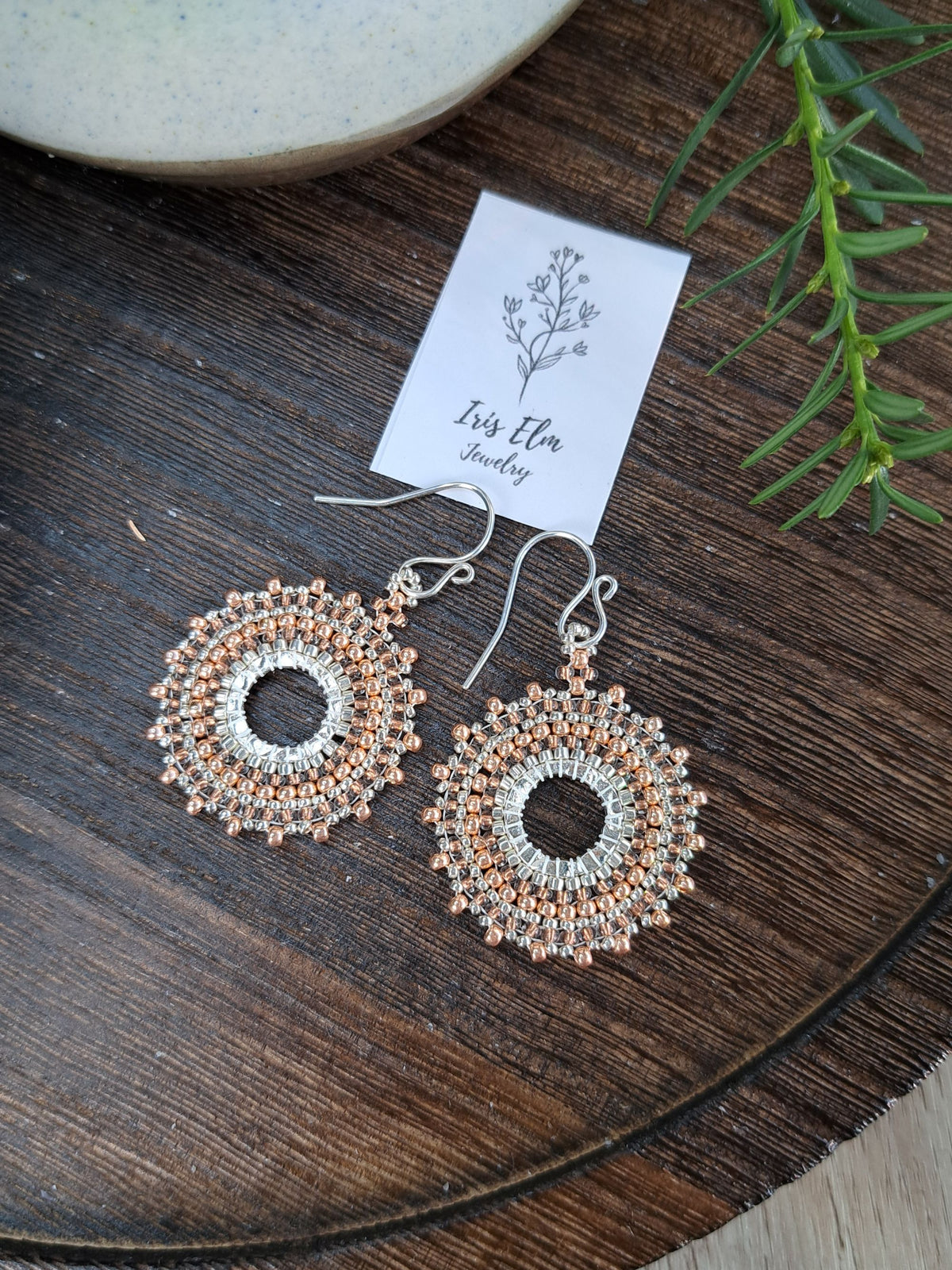 a pair of handmade beadwork circular hoop earrings woven with silver and copper colored glass seed beads by Iris Elm Jewelry on wooden surface