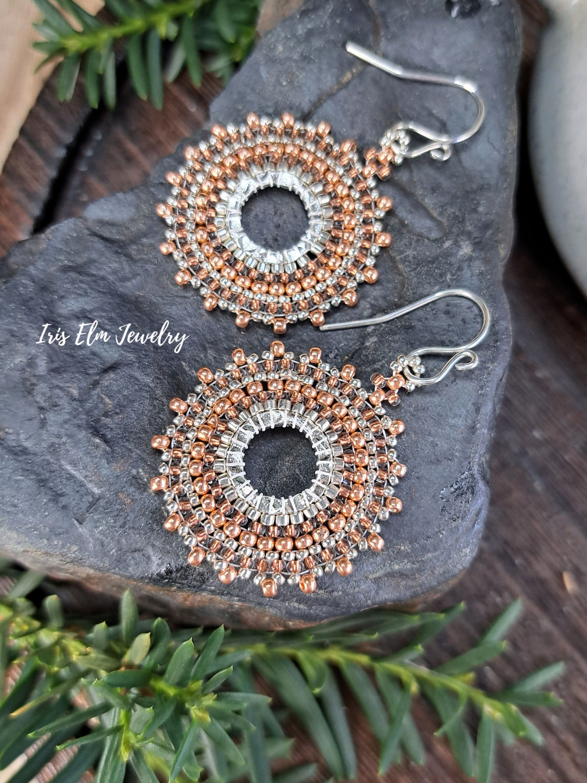 a pair of handmade beadwork circular hoop earrings woven with silver and copper colored glass seed beads by Iris Elm Jewelry on rocky surface