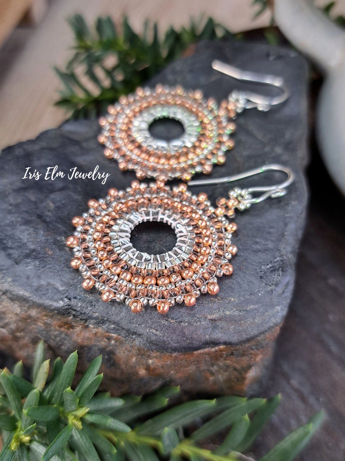 a pair of handmade beadwork circular hoop earrings woven with silver and copper colored glass seed beads by Iris Elm Jewelry on rocky surface