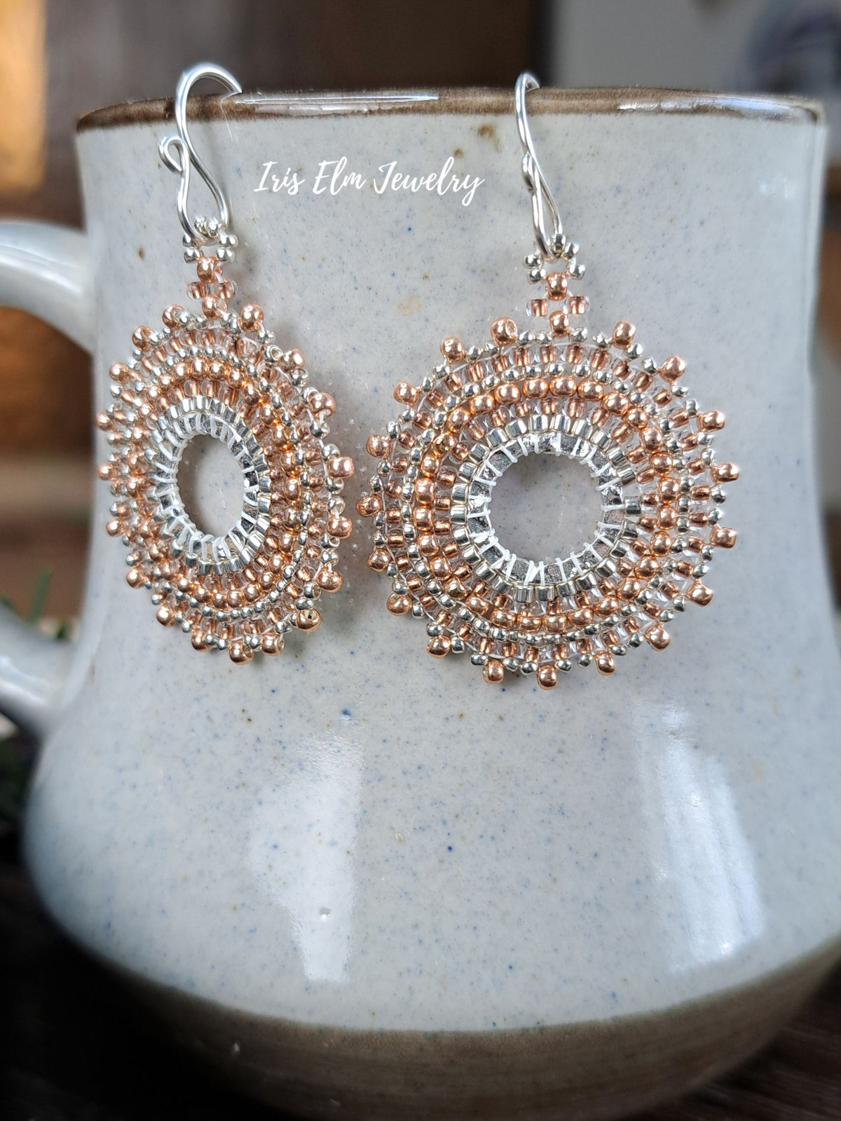 a pair of handmade beadwork circular hoop earrings woven with silver and copper colored glass seed beads by Iris Elm Jewelry on ceramic mug