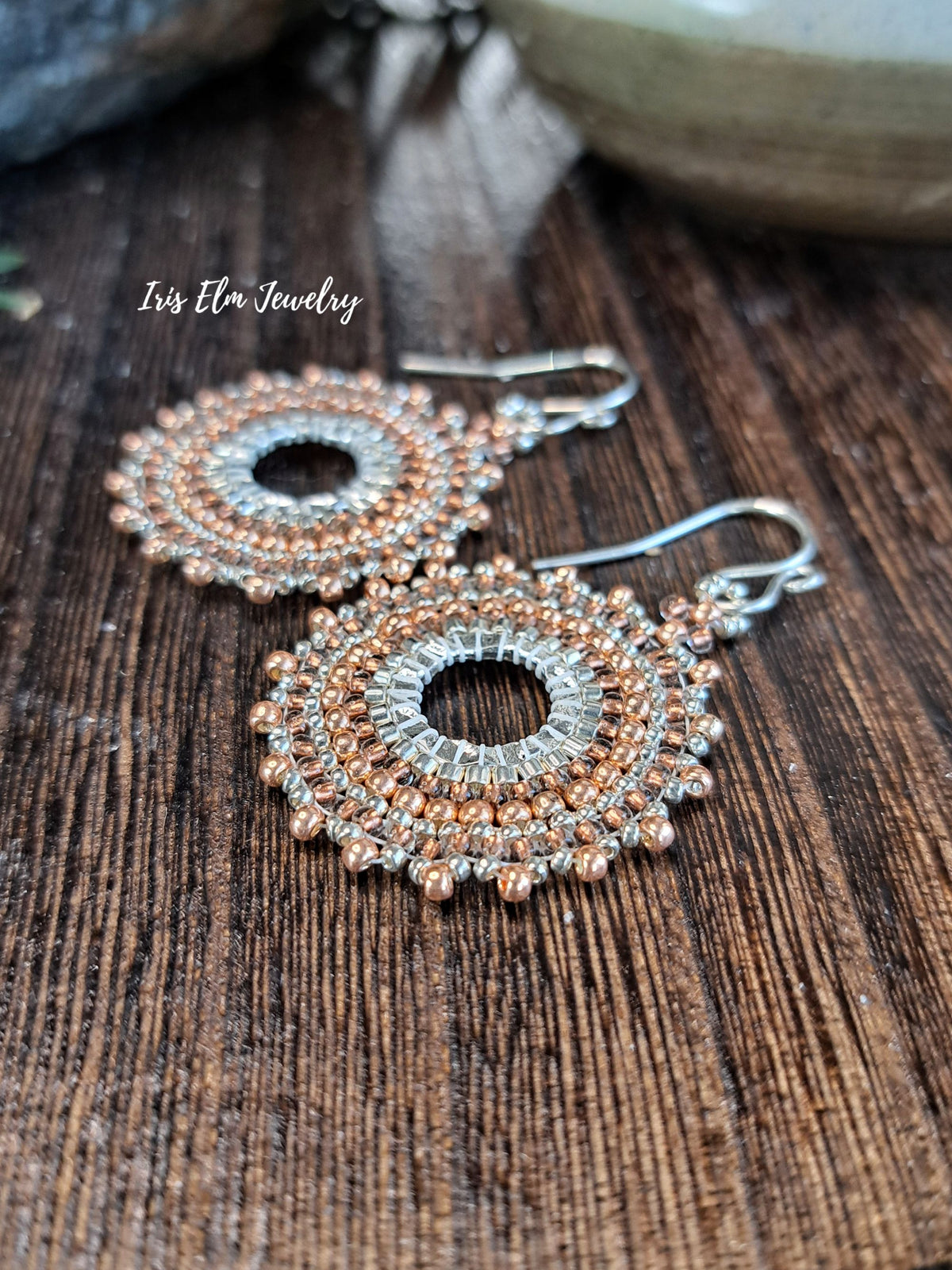 a pair of handmade beadwork circular hoop earrings woven with silver and copper colored glass seed beads by Iris Elm Jewelry on wooden surface