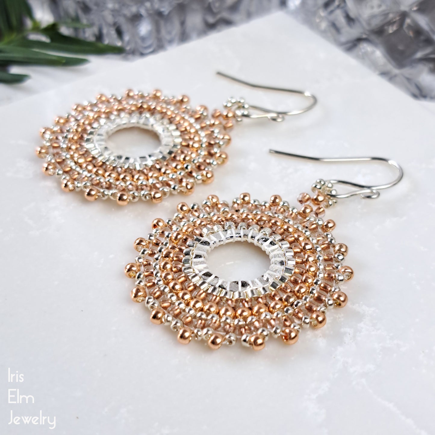 Pair of beaded copper and silver circular handmade hoop earrings on a marble surface with 'Iris Elm Jewelry' branding.