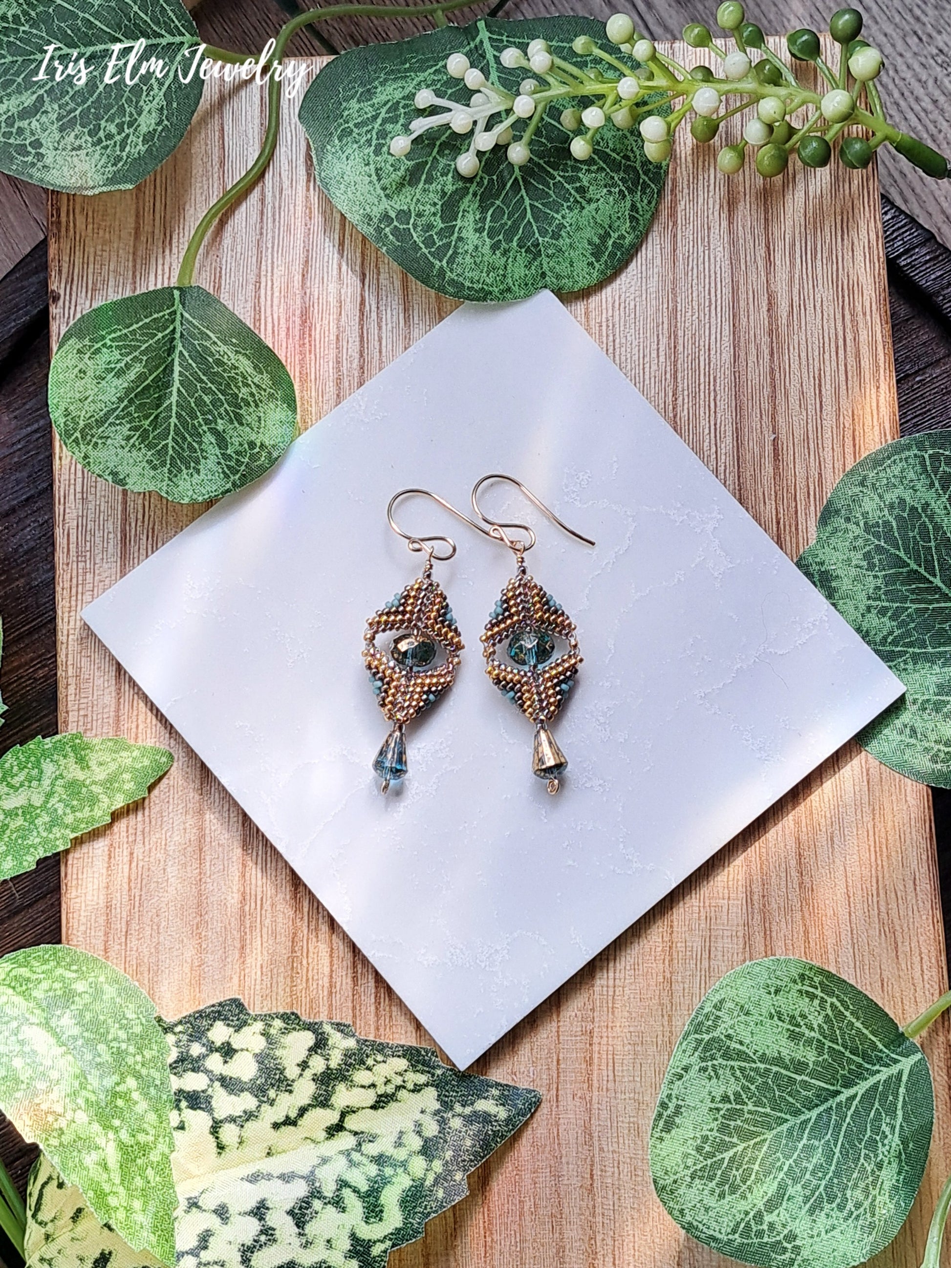 Pair of beadwork gold and aqua art deco style glass earrings on a white square with green leaves around on a wooden surface