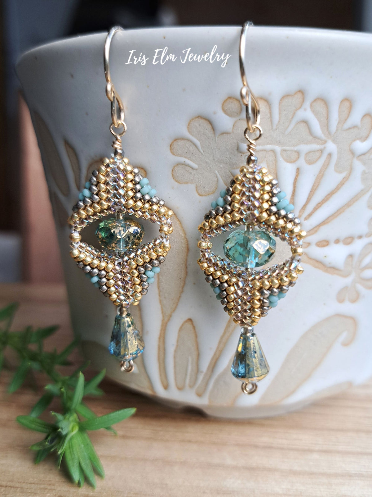 A pair of handmade beaded drop earrings with gold-toned metal, turquoise beads, and green faceted crystals, hanging in front of a white ceramic cup on a wooden surface.