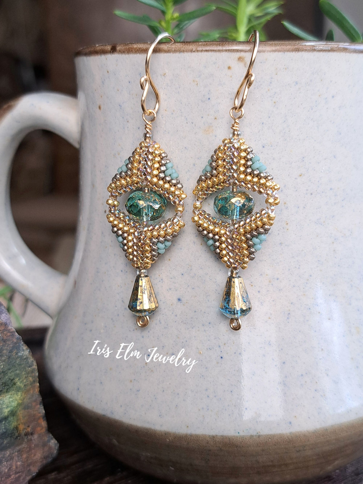A pair of handmade bohemian-style dangle earrings with gold seed beads, turquoise beads, and green faceted crystals, hanging in front of a ceramic mug. The brand name 'Iris Elm Jewelry' is visible as a watermark on the mug.