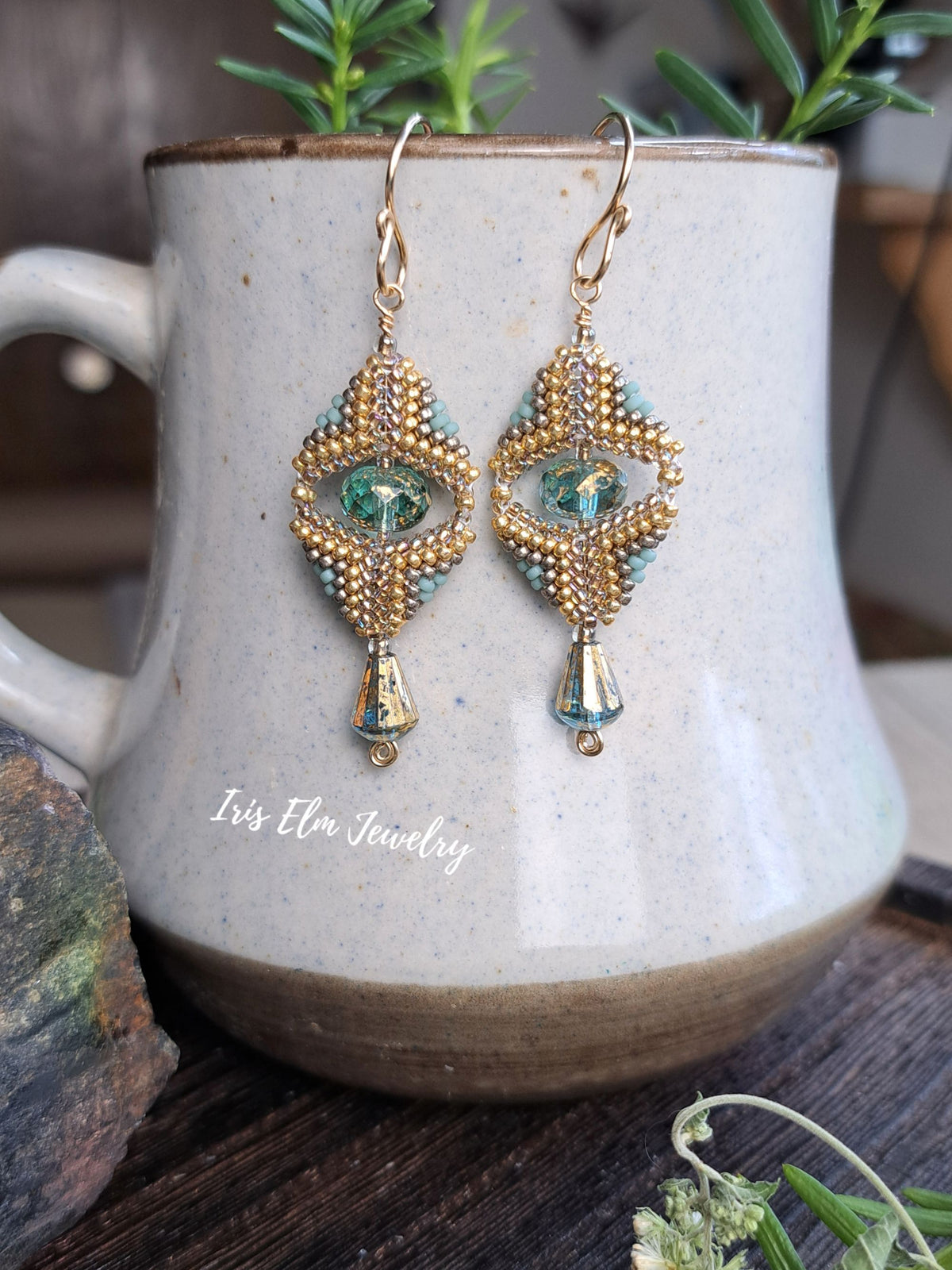 A pair of gold and turquoise beaded drop earrings displayed on a speckled ceramic mug. The earrings feature intricate gold and turquoise beadwork with a central green teardrop glass bead and dangling gold beads.
