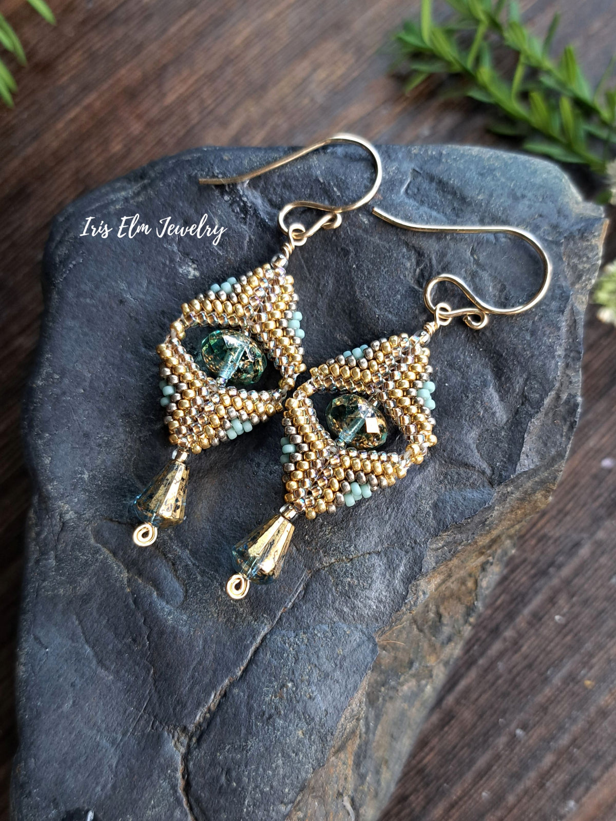 A pair of handmade beaded dangle earrings with gold-tone hooks and frames, decorated with small gold and light blue beads and a central green faceted glass bead. They are displayed on a dark slate rock with a wooden surface and green leaves in the background.