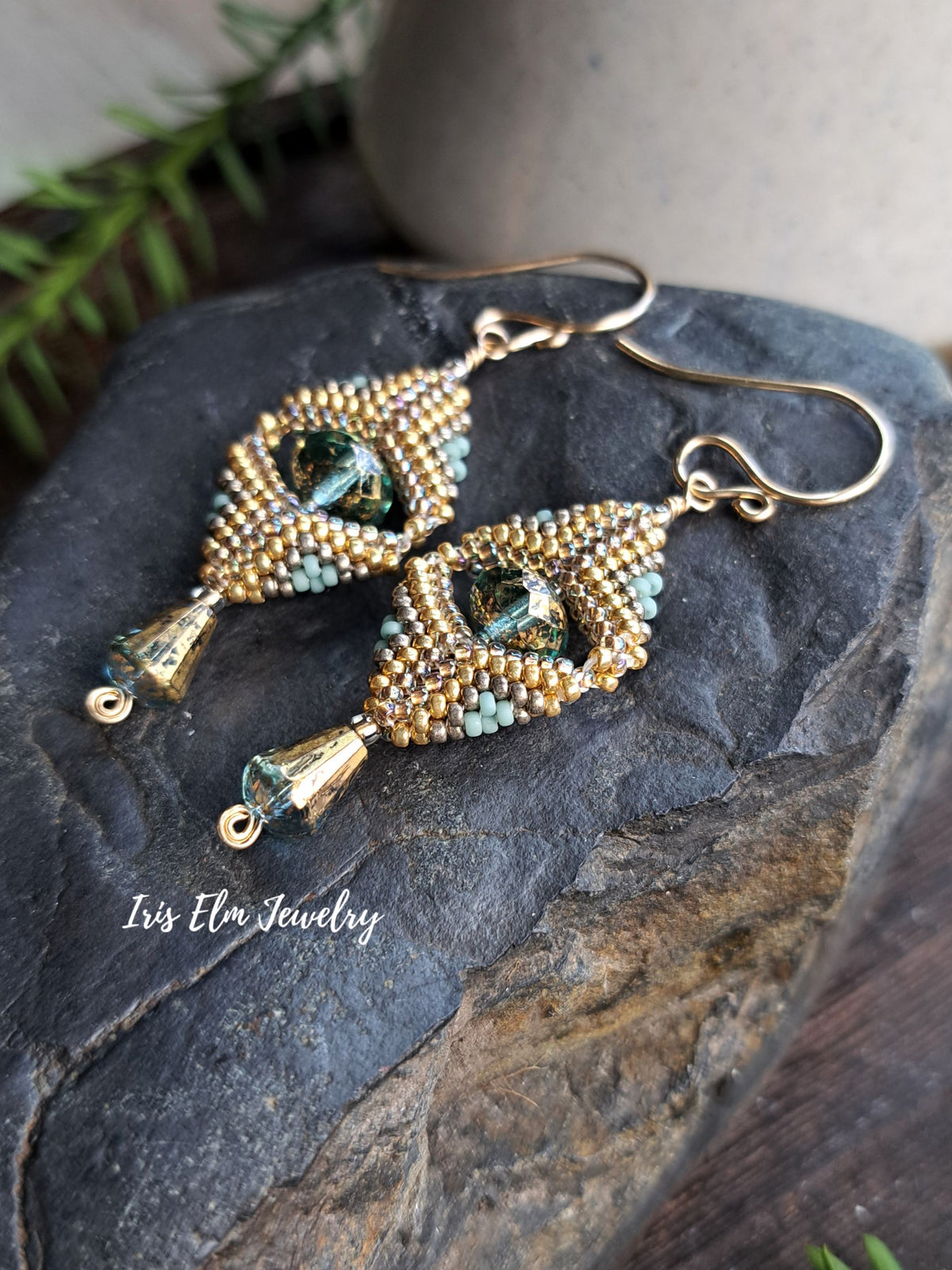 A pair of handmade beaded dangle earrings with gold-tone hooks and frames, decorated with gold, light blue, and green beads, resting on a dark textured stone. The brand name 'Iris Elm Jewelry' is watermarked on the lower left.