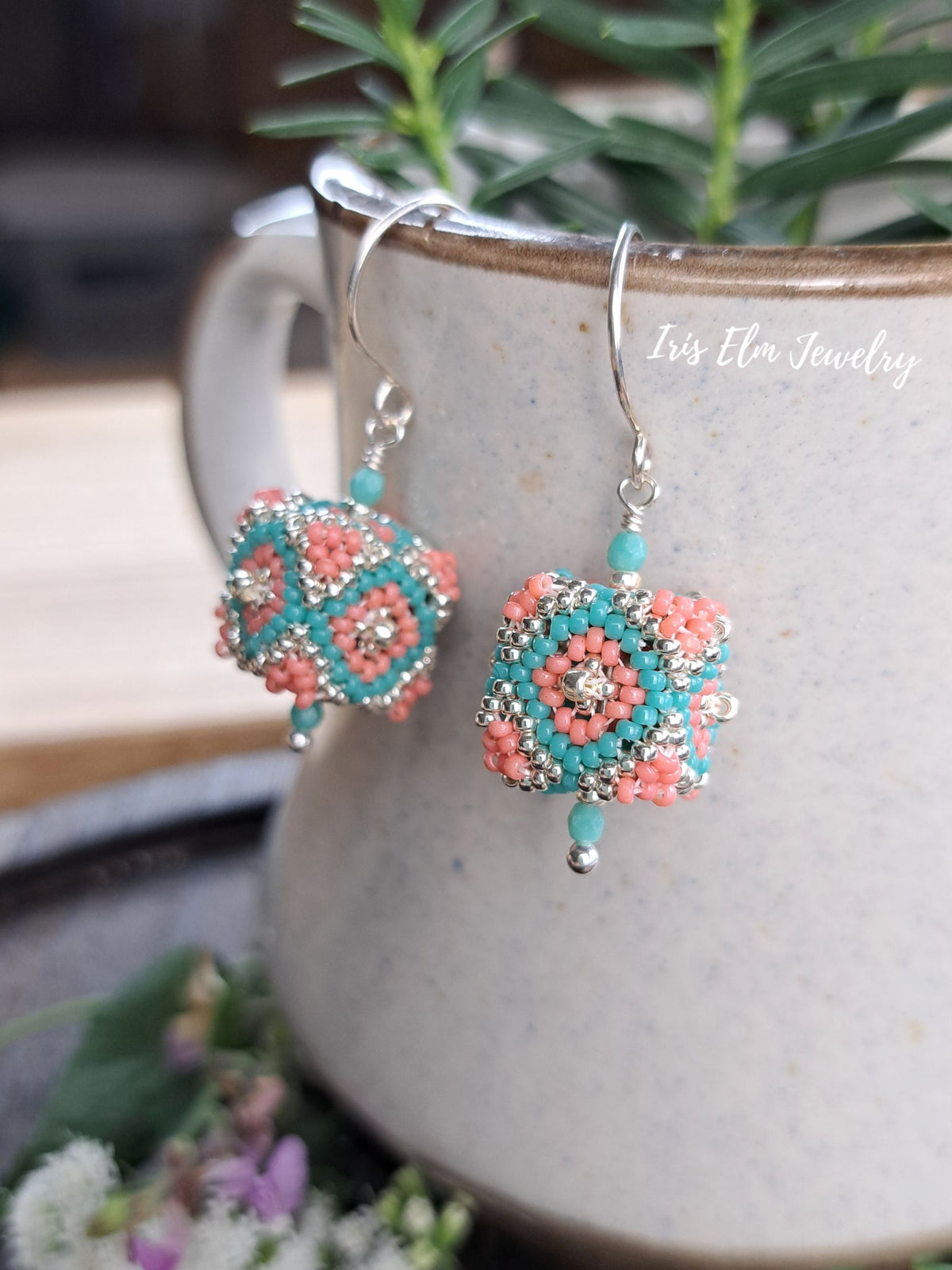 A pair of handmade cube seed bead beaded dangle earrings with turquoise and coral geometric patterns, hanging from silver hooks in front of a ceramic mug with green leaves in the background. The brand name 'Iris Elm Jewelry' is watermarked on the right.