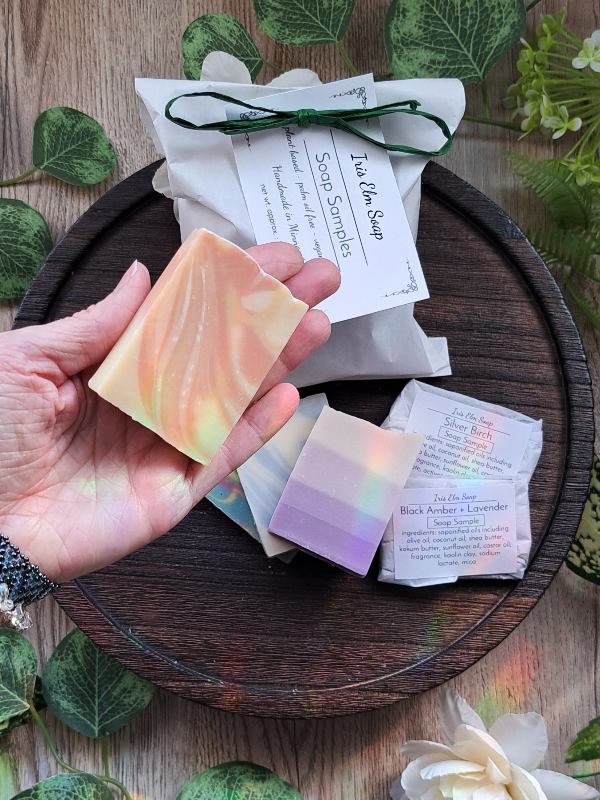 Hand holding a handmade cold process vegan soap bar with Iris Elm Soap samples on a wooden tray.