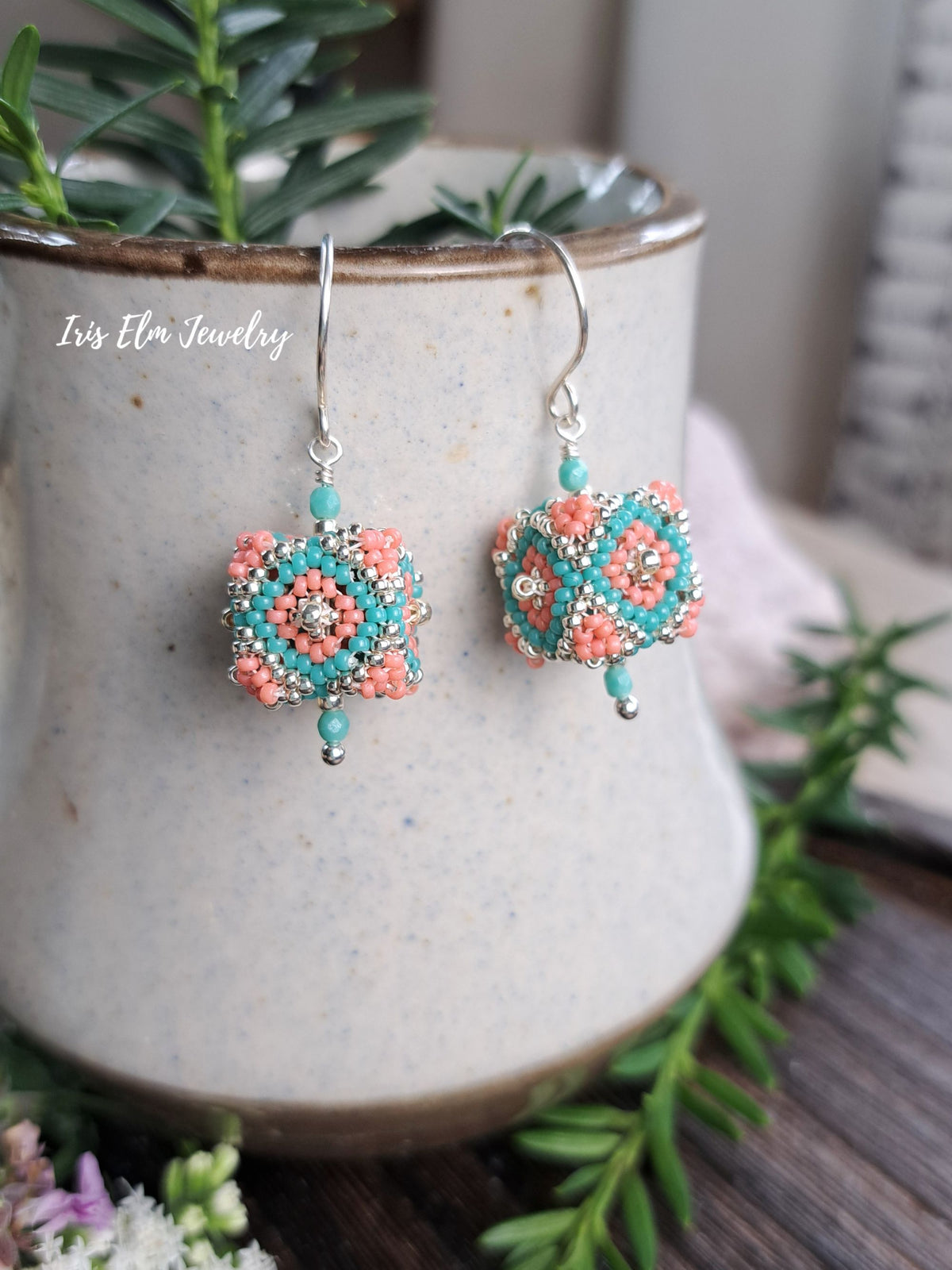 A pair of handmade beaded drop earrings by Iris Elm Jewelry, featuring turquoise and coral beads with a silver hook. The earrings are displayed on a speckled ceramic vase with green foliage in the background.