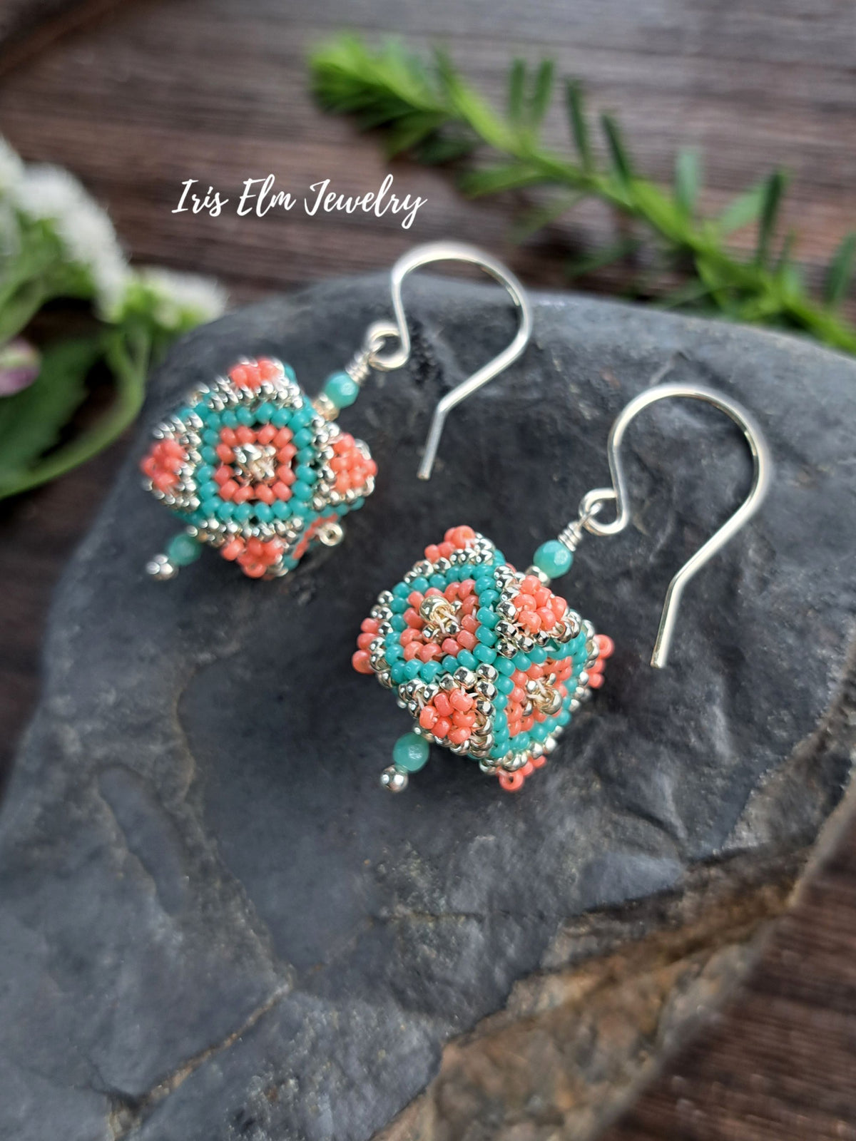 A pair of handmade beaded dangle earrings with coral and turquoise geometric patterns, silver-toned metal hooks, and small silver bead drops, displayed on a dark slate stone with green foliage in the background. The text 'Iris Elm Jewelry' is visible in the upper left.