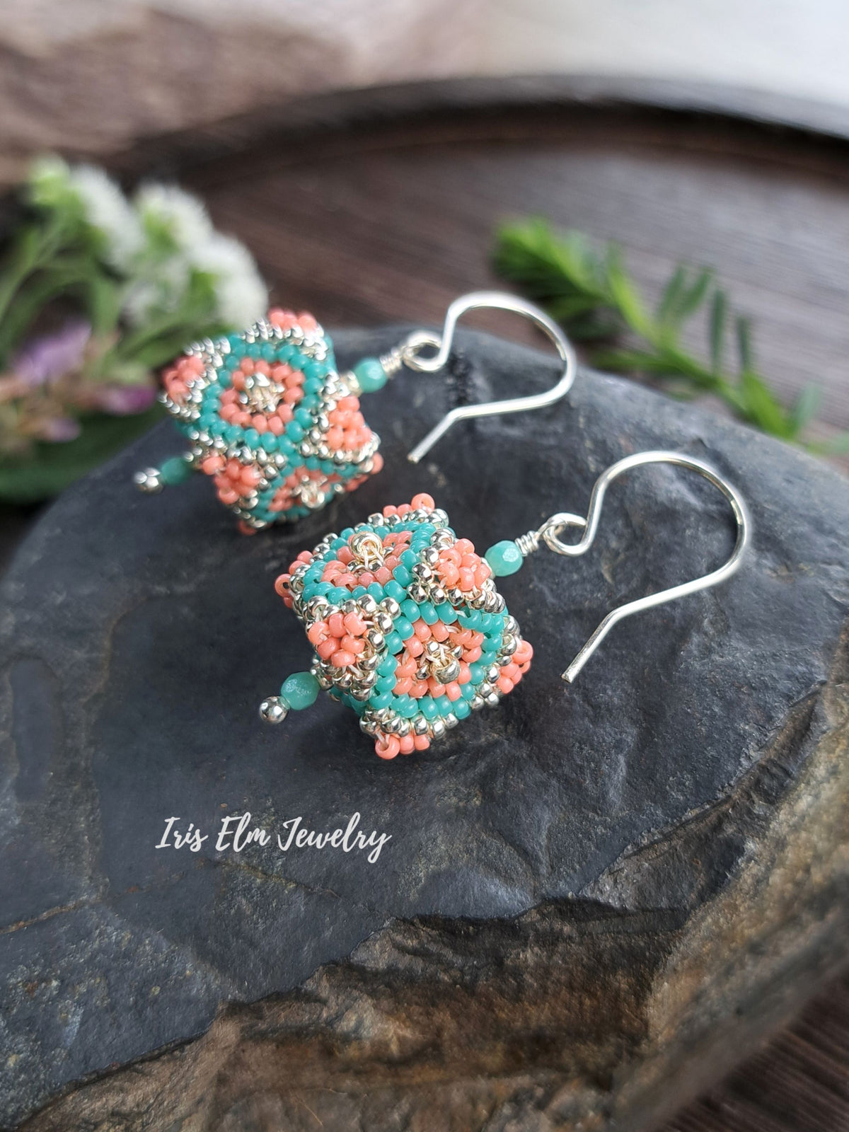 Two handmade beaded dangle earrings with coral pink and turquoise geometric patterns on silver hooks, displayed on a dark slate stone with a 'Iris Elm Jewelry' watermark.