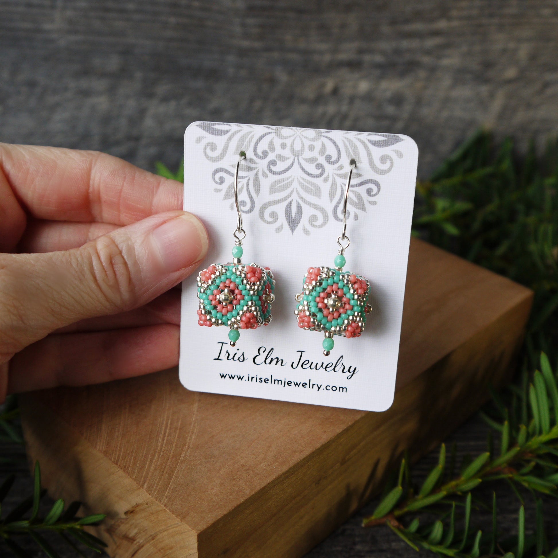Pair of seed bead woven cube southwestern earrings on a 'Iris Elm Jewelry' card held by a hand with a natural background.
