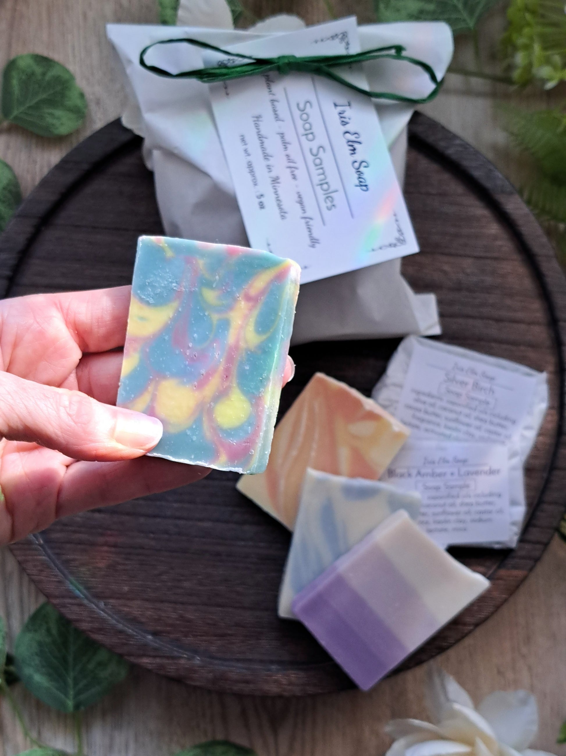 Hand holding a colorful soap bar with more variety sample soaps and packaging on a wooden surface.