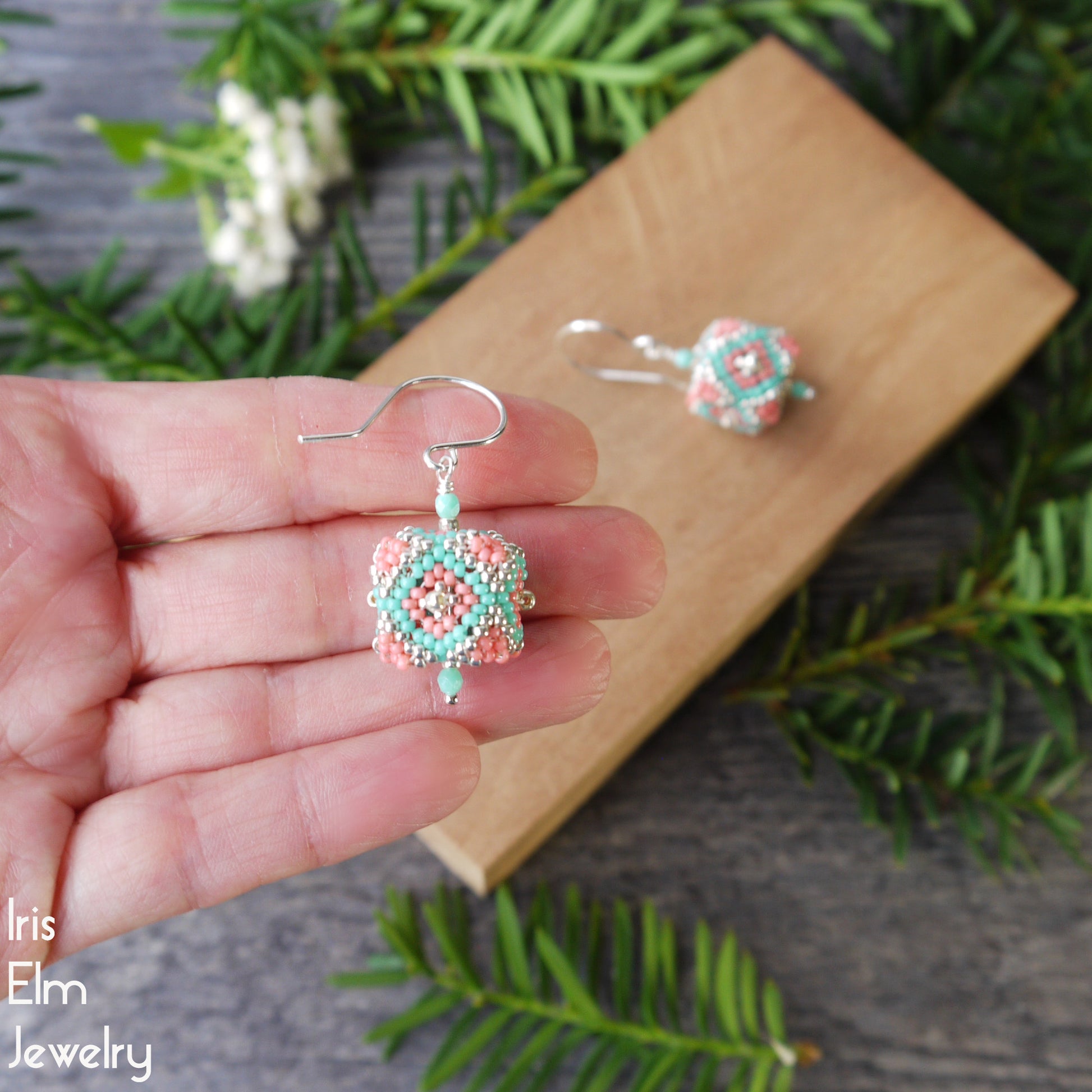 Beaded cube shaped turquoise and coral pink glass seed bead earrings held in a hand with a wooden box and greenery in the background, featuring 'Iris Elm Jewelry'.