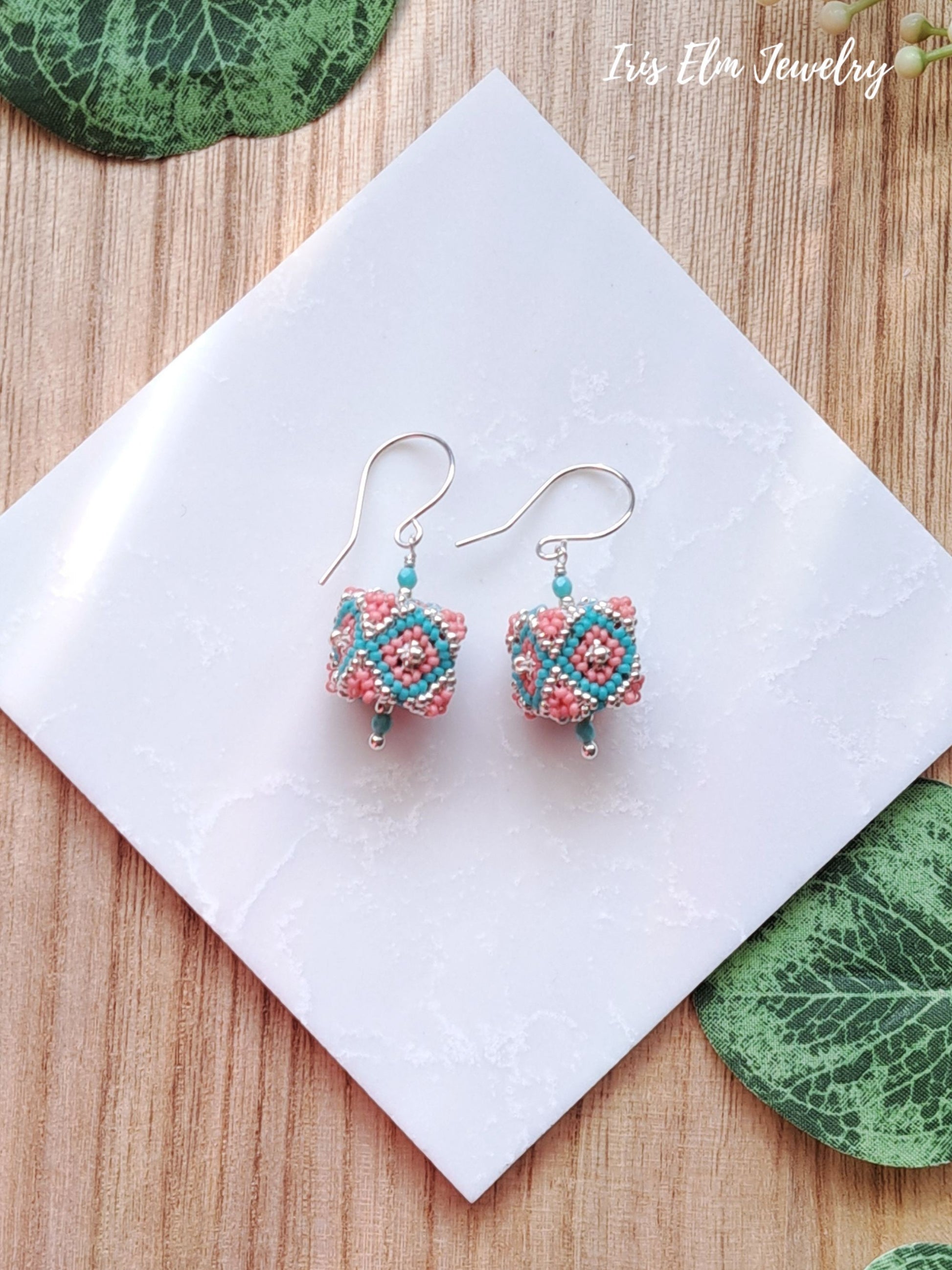 Pair of beaded coral pink and turquoise seed bead cube earrings on a white napkin with 'Iris Elm Jewelry' branding.