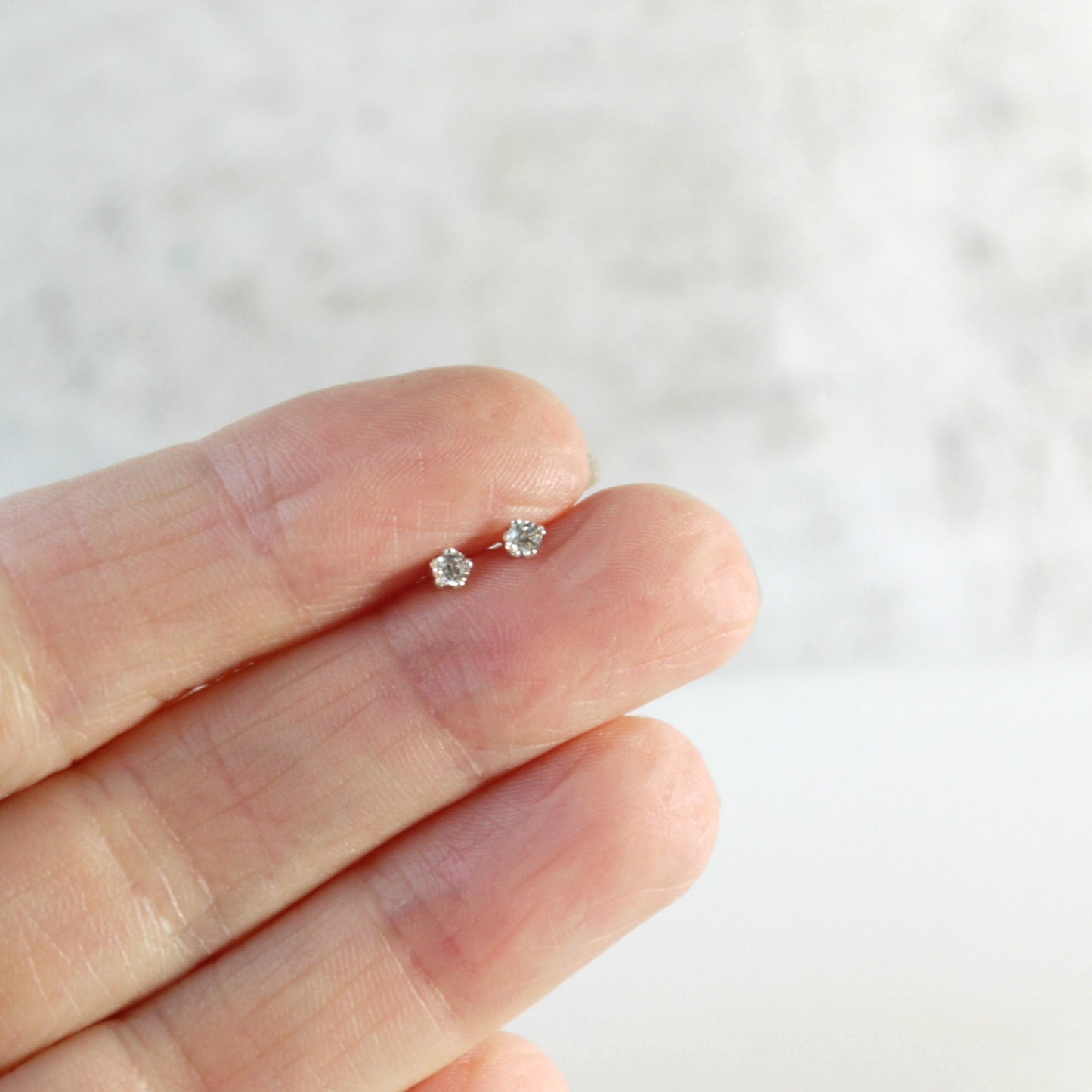 tiny 2mm clear cubic zirconia sterling silver stud earrings held between fingers against a neutral background
