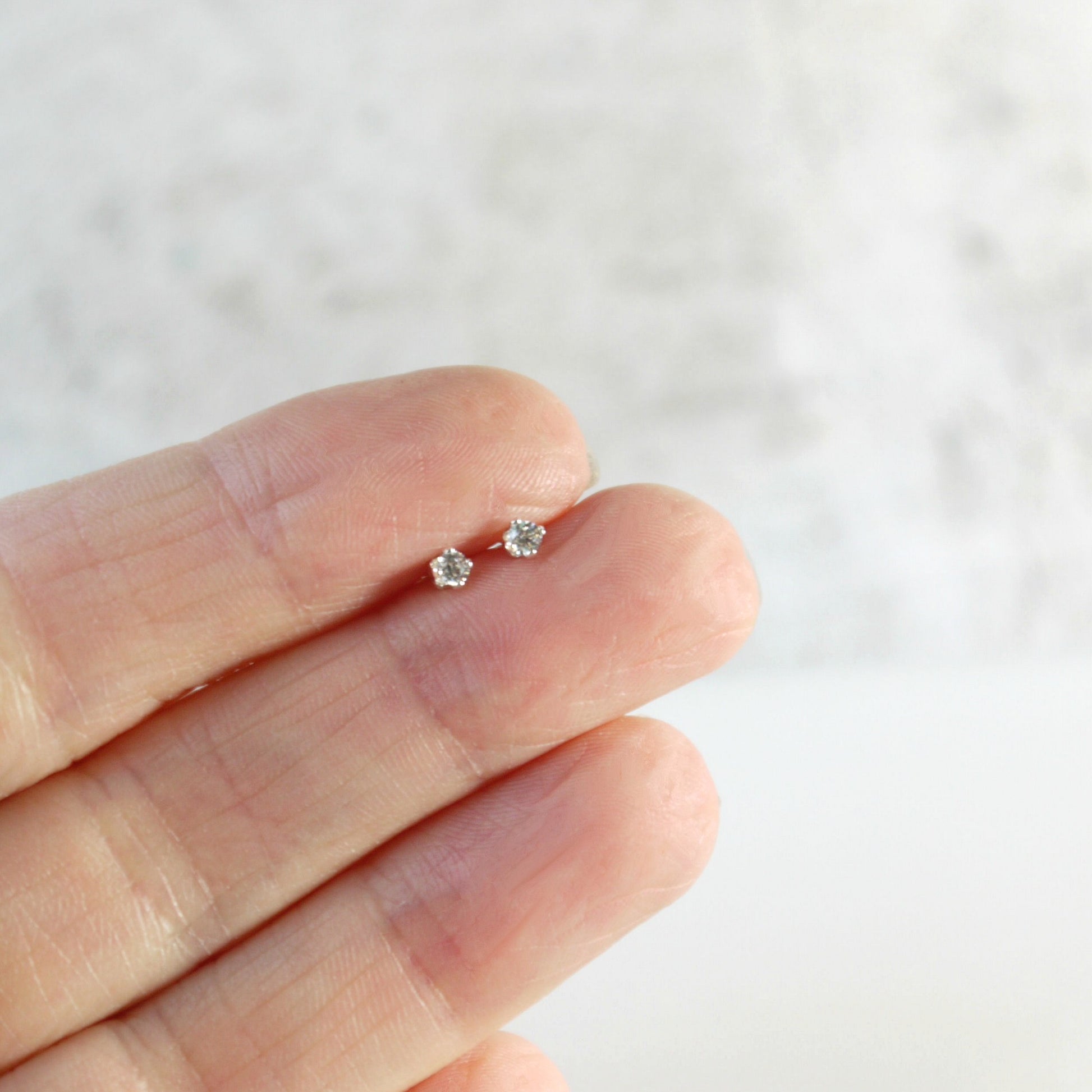 tiny 2mm clear cubic zirconia sterling silver stud earrings held between fingers against a neutral background