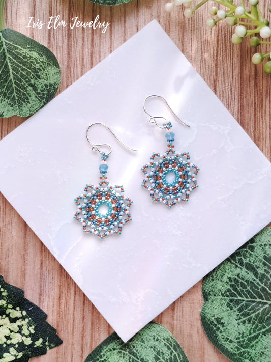 Beaded blue apatite white orange seed bead woven mandala earrings on a white tile with green leaves in the background