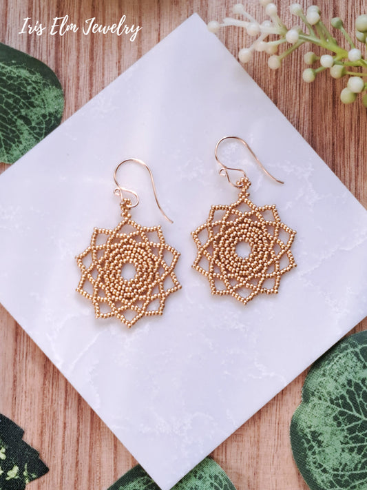 Gold seed bead woven mandala earrings on a white napkin with 'Iris Elm Jewelry' branding, surrounded by green leaves and flowers.