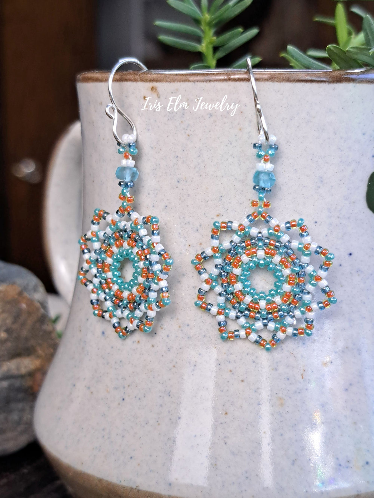 A pair of handmade beaded dangle earrings with silver fishhook wires, featuring intricate geometric patterns in turquoise, white, and orange. The earrings are displayed hanging from a speckled ceramic mug, with a soft-focus background showing green leaves and a wooden surface.