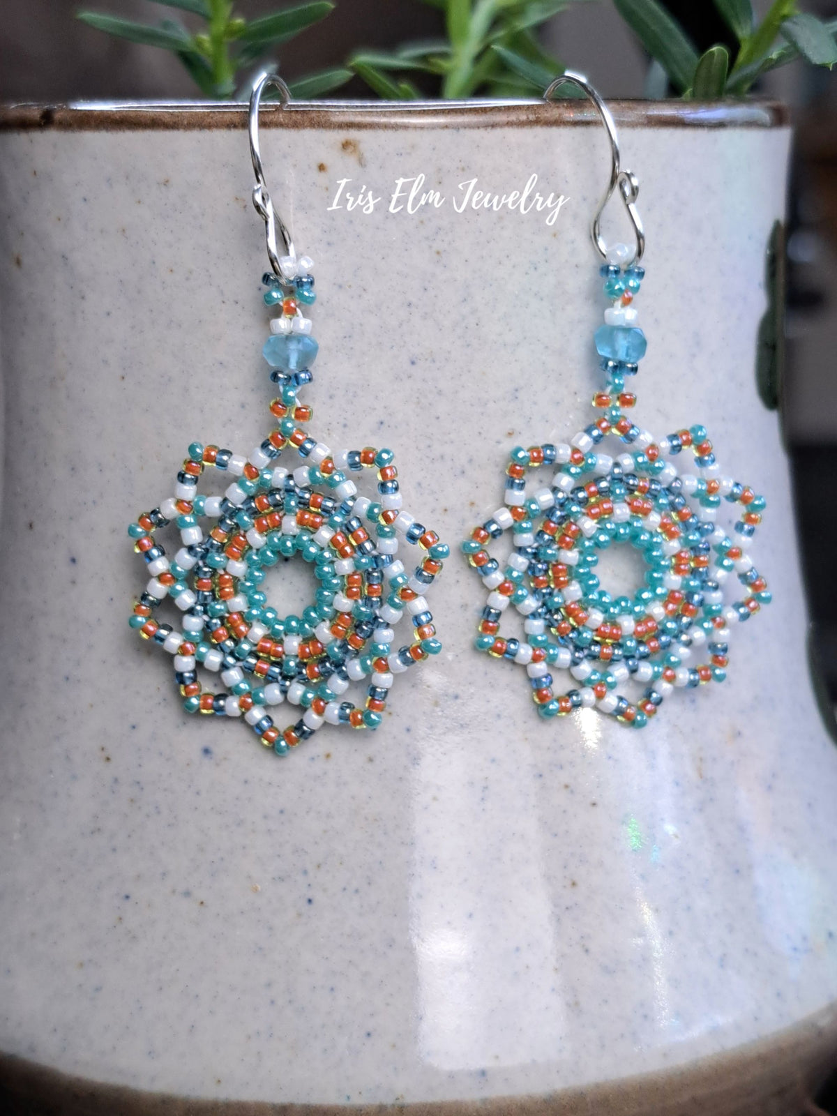 A pair of handmade beaded dangle earrings with silver fishhook wires, featuring intricate geometric patterns in turquoise, white, and orange. The earrings are displayed hanging from a speckled ceramic mug, with a soft-focus background showing green leaves and a wooden surface.