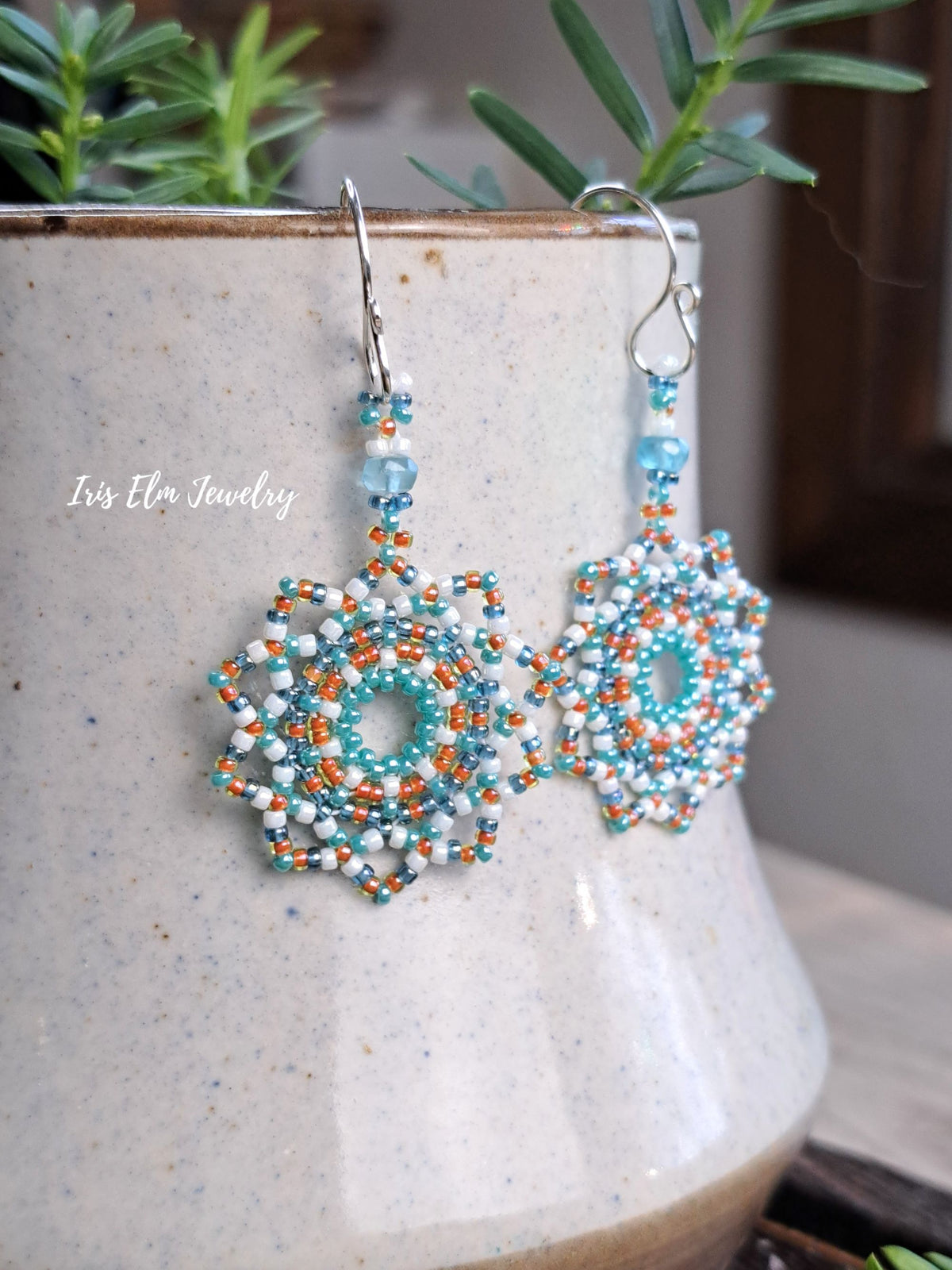 A pair of handmade beaded dangle earrings with intricate snowflake-like floral patterns in turquoise, white, and orange, hanging from silver fishhook wires. The earrings are displayed in front of a speckled ceramic vase with green leaves in the background.