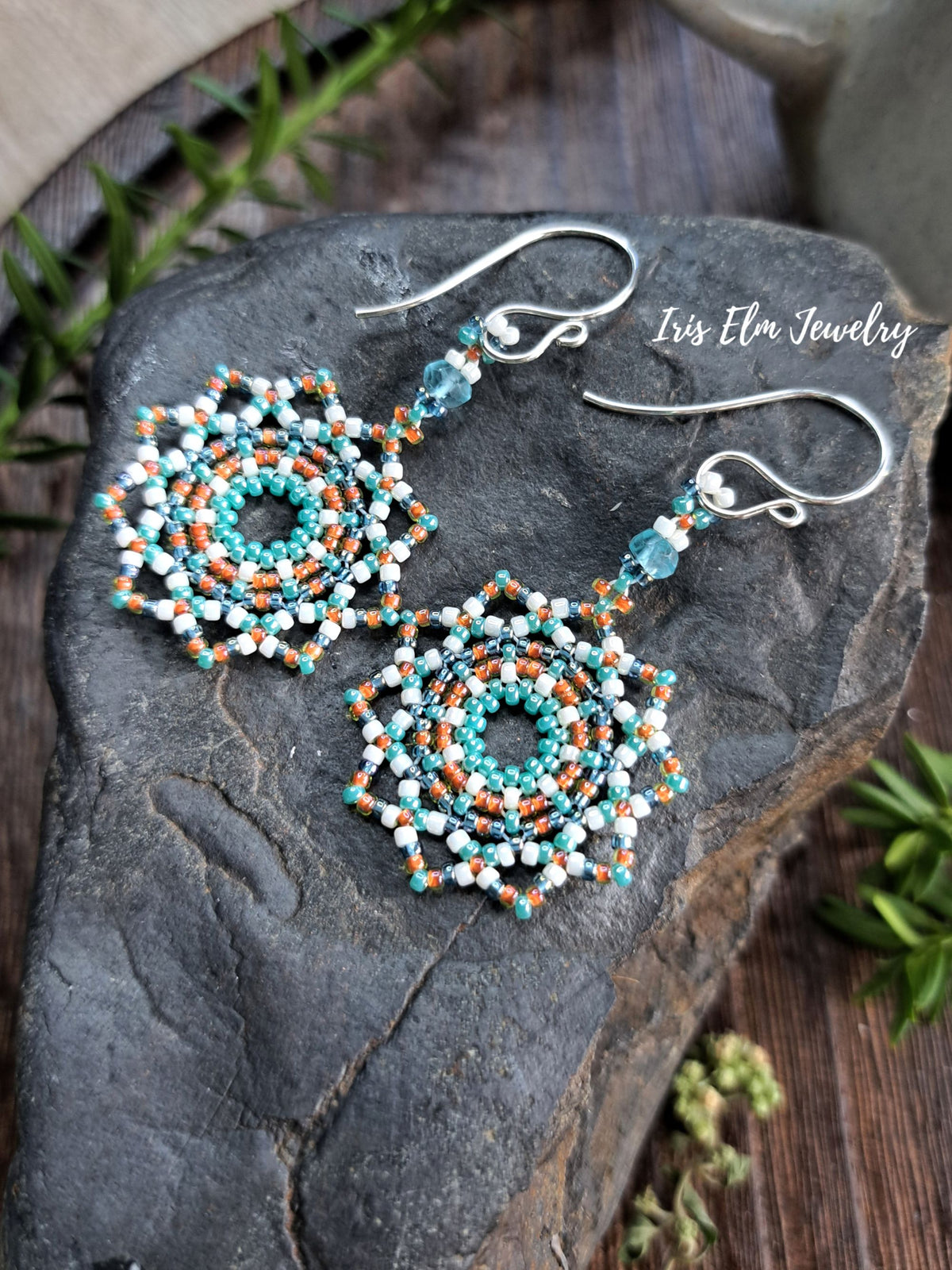 A pair of handmade beaded dangle earrings with silver fishhook wires, displayed on a dark textured stone. The earrings feature intricate circular mandala patterns in turquoise, white, and orange, with small faceted light blue beads hanging from the hooks.