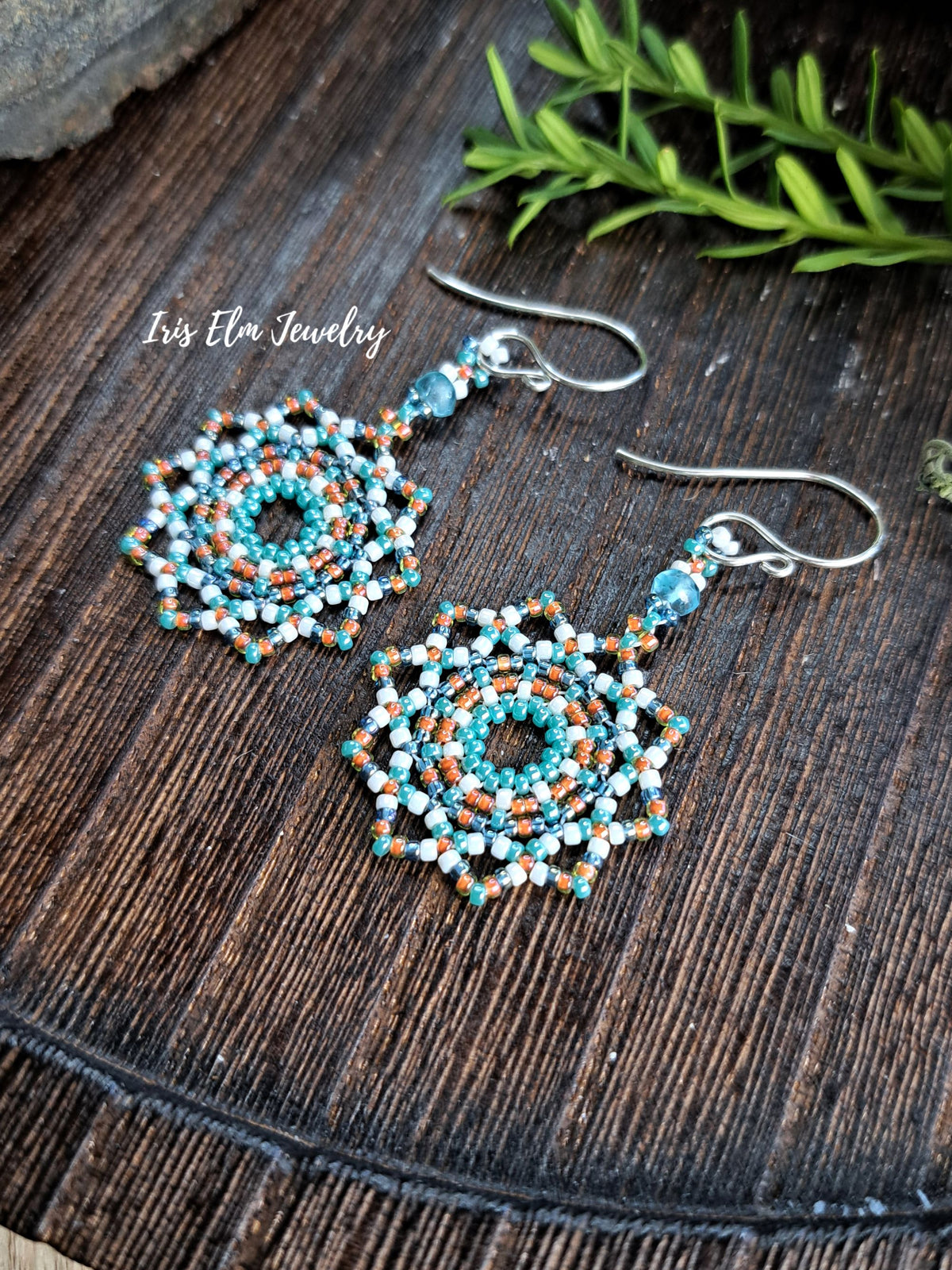 A pair of handmade beaded dangle earrings with intricate circular patterns in turquoise, white, and orange, hanging from silver fishhook wires on a dark wooden surface with green leaves in the background. The text 'Iris Elm Jewelry' is visible on the left.