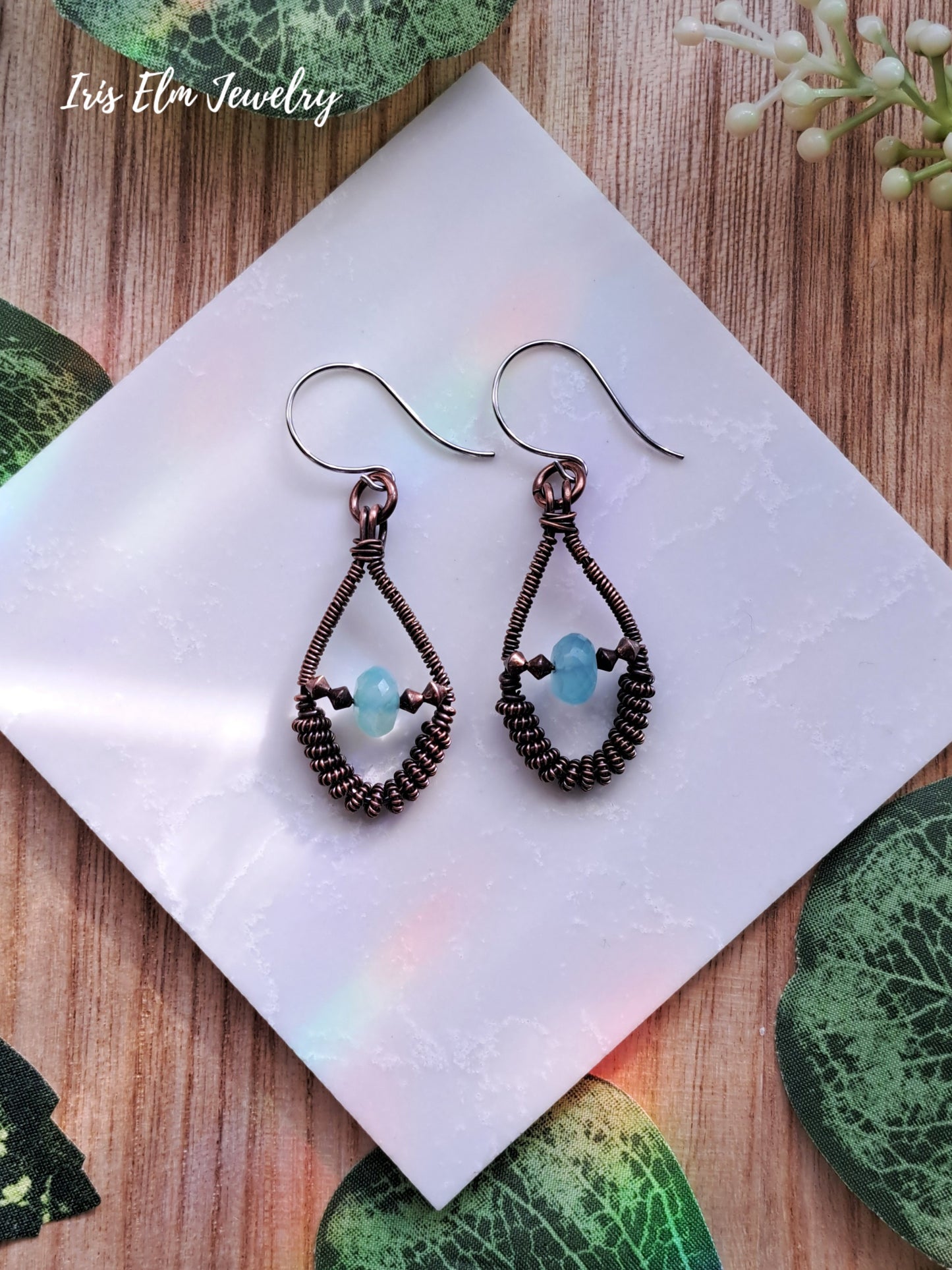 Pair of wire-wrapped earrings with blue chalcedony beads on a white square card, surrounded by green leafy elements.